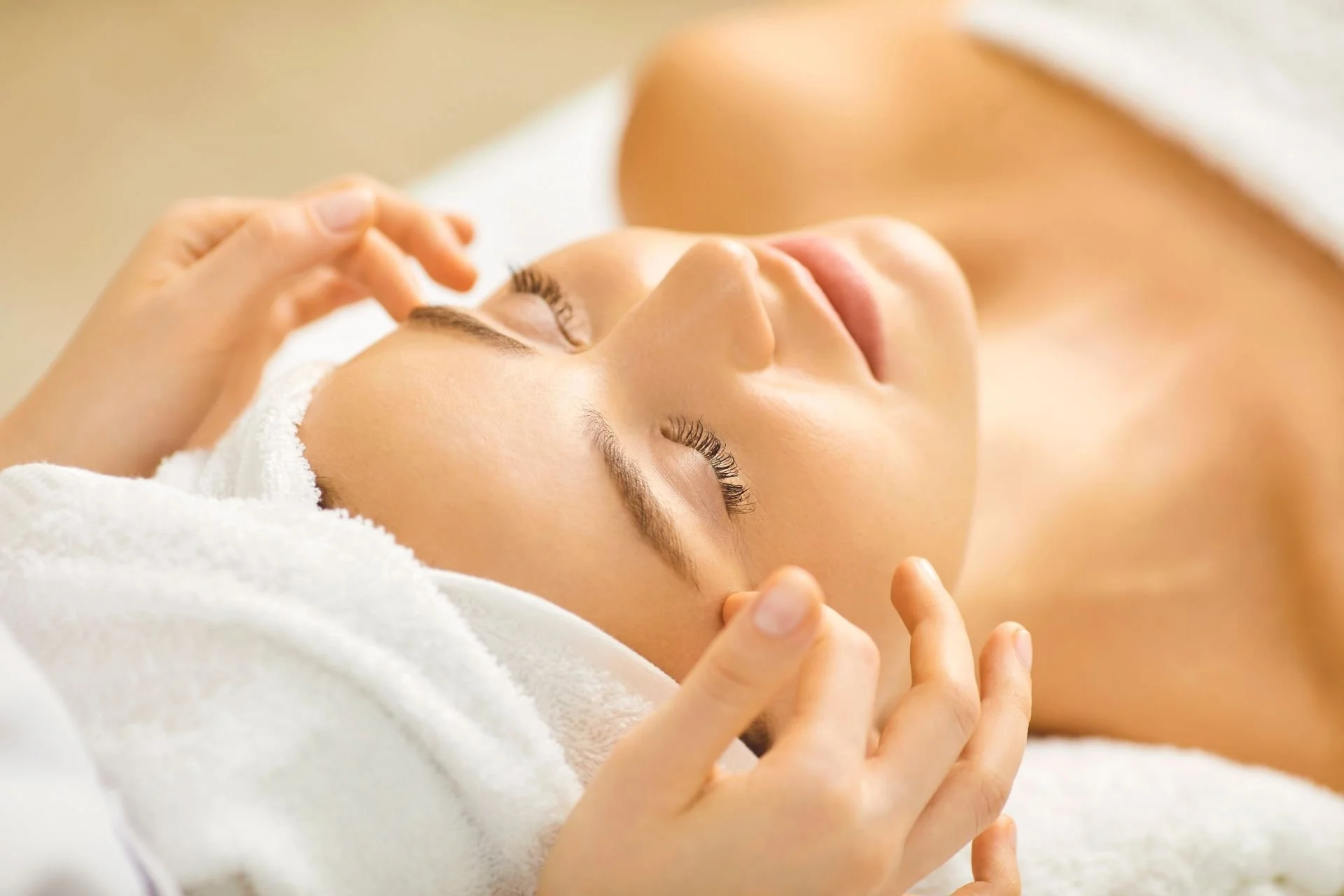 A woman receives a facial treatment, lying on a towel with eyes closed, while a cosmetologist gently massages her face.
