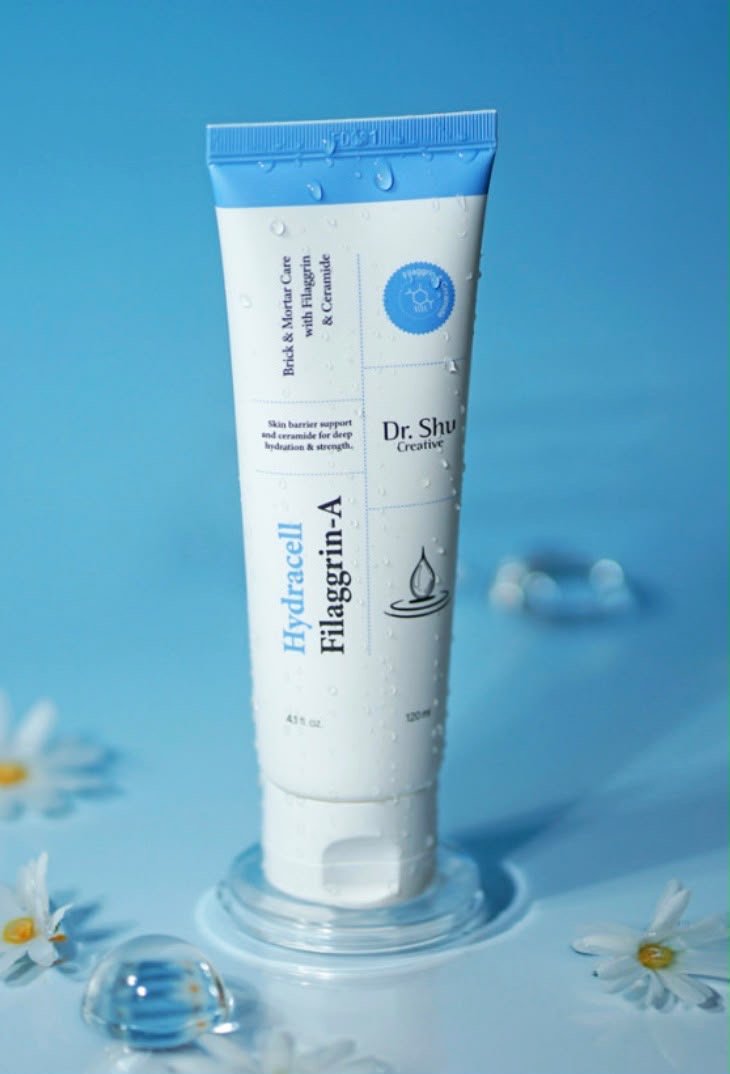 A white tube of Dr. Shulz Hydracell Filaggrin-A skincare product with water droplets on it, standing on a clear stand in a blue background, surrounded by small daisies and water bubbles.