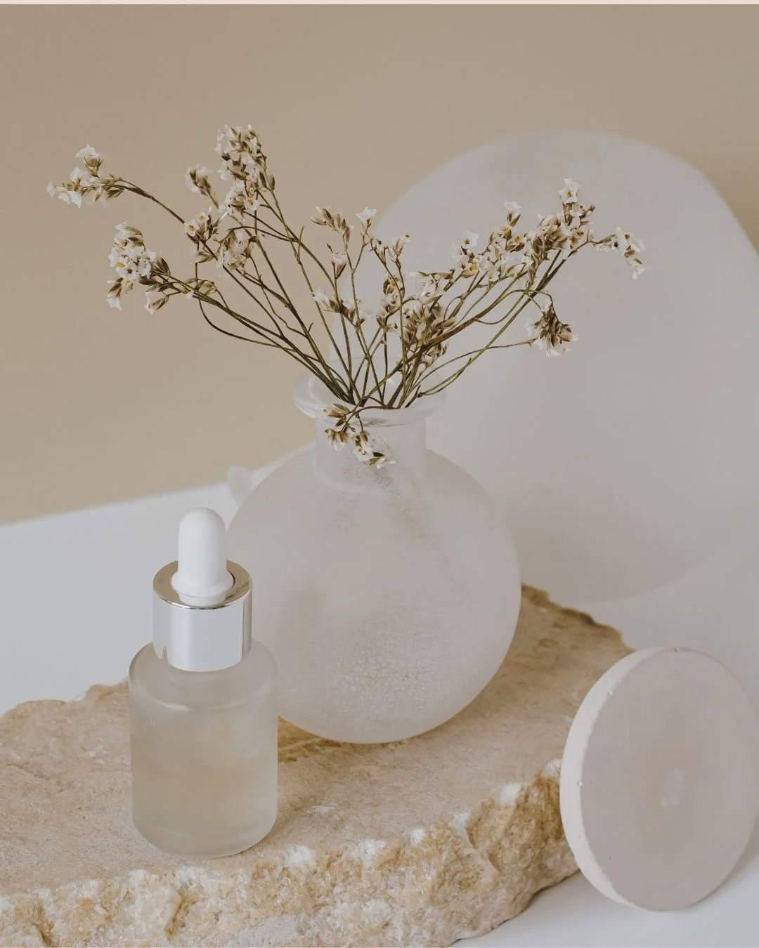 A minimalist still life featuring a frosted glass vase with dried white flowers, a small frosted dropper bottle, and a round frosted glass object on a beige stone surface against a neutral background.