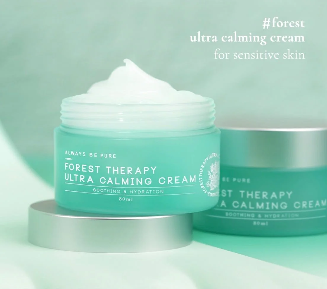 A jar of Forest Therapy Ultra Calming Cream with the lid off, showing the white cream inside, placed on a silver, round platform against a soft green background.
