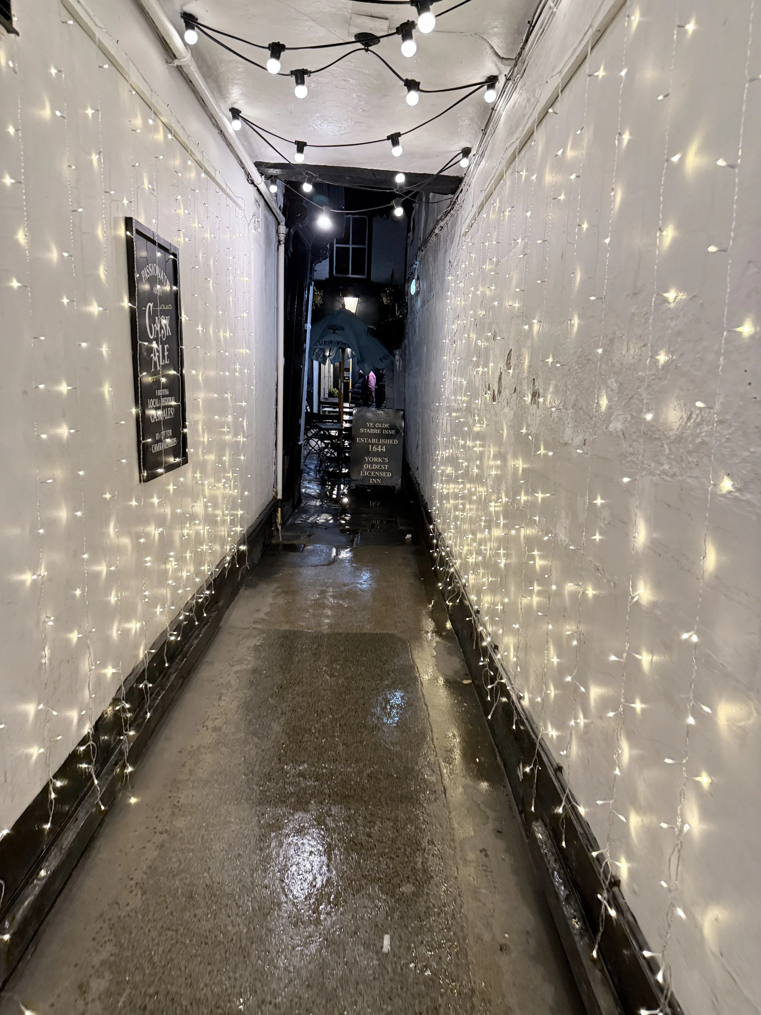 A narrow alleyway decorated with white string lights on both sides and ceiling, leading to an outdoor seating area with tables and an umbrella, wet pavement reflecting the lights.