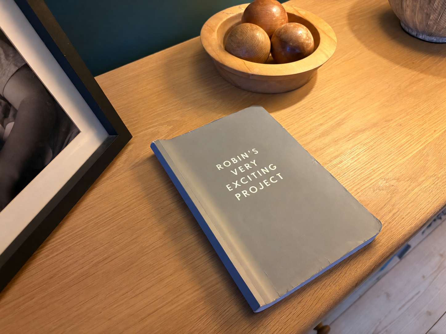 A grey notebook rests on a wooden counter top, next to a photo frame and a wooden bowl. The title, 'Robin's Very Exciting Project', is printed in capital letters on the cover of the notebook.