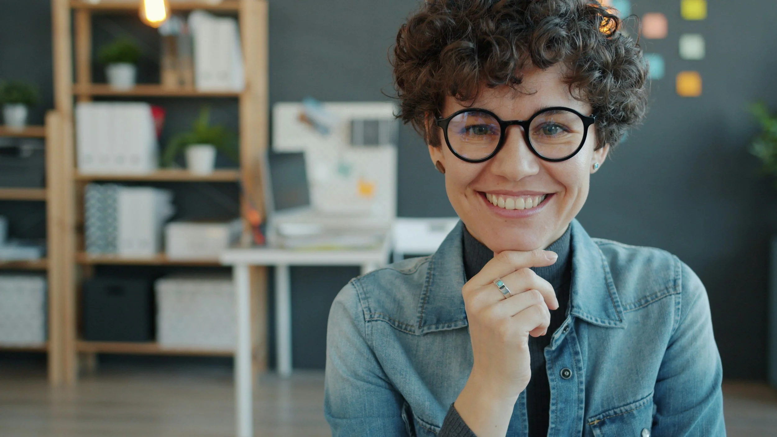A lady with brown curly hair, black circular glasses, and wearing a light blue denim jacket over a dark blue roll neck top smiles directly at the camera with the thumb and forefinger of her right hand holding her chin.
