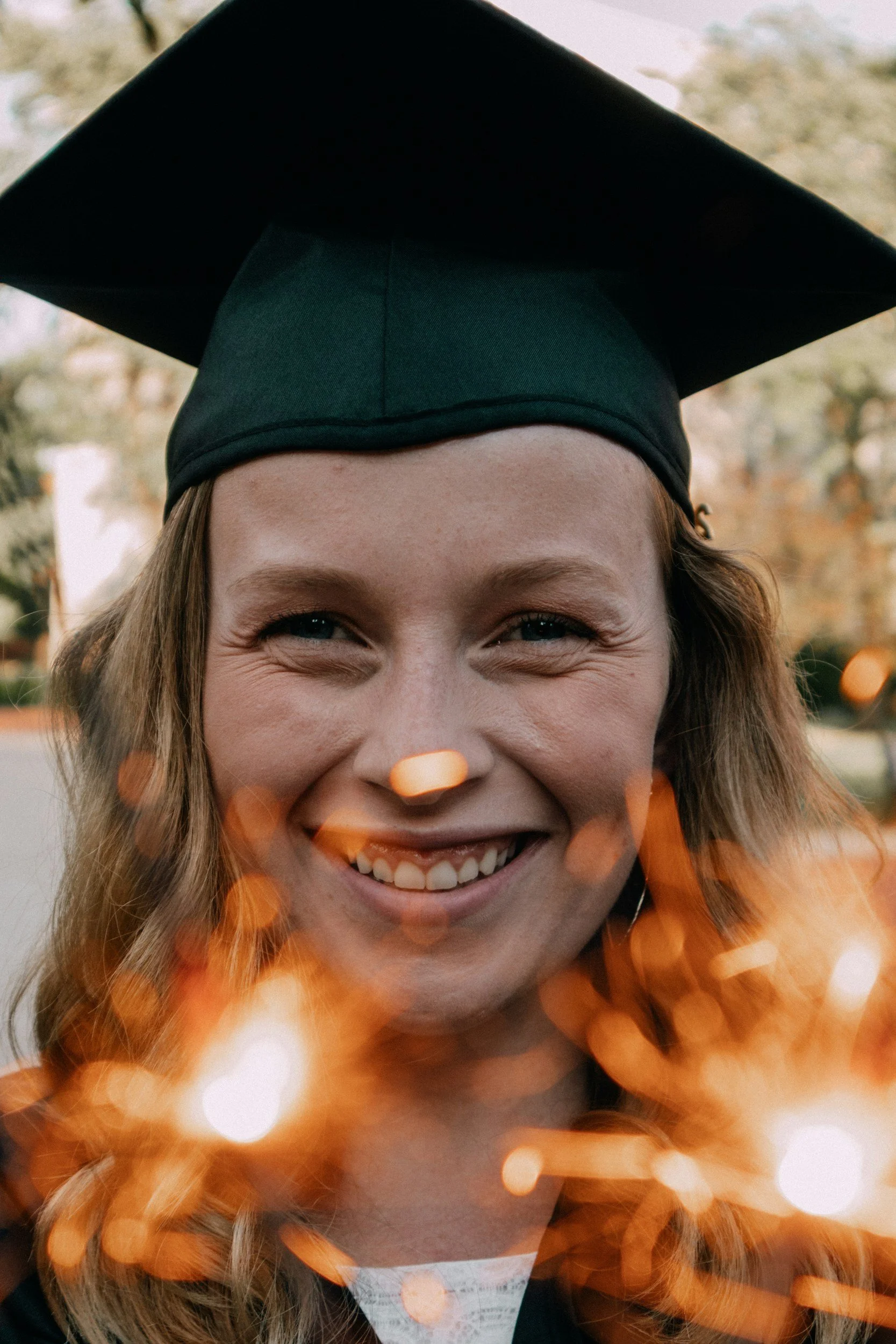 A headshot of blonde-haired female student wearing a mortarboard. She is facing the camera smiling, and she appears to be holding two lit sparklers, with the sparkles visible but blurred in the lower foreground of the image.