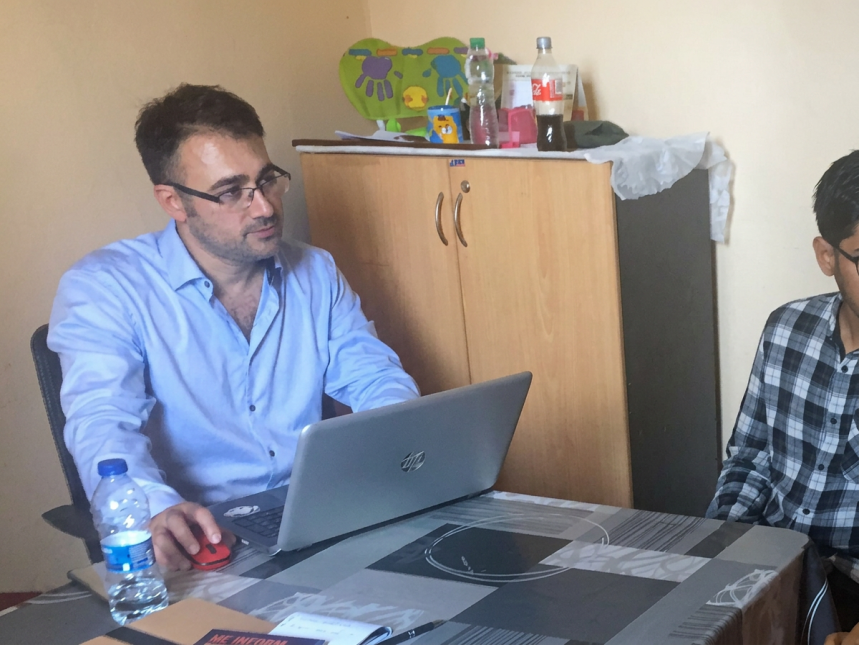A man with glasses and a light blue button-up shirt, identified as Robin Hodges, sits at a patterned table using a silver HP laptop to conduct a needs assessment..