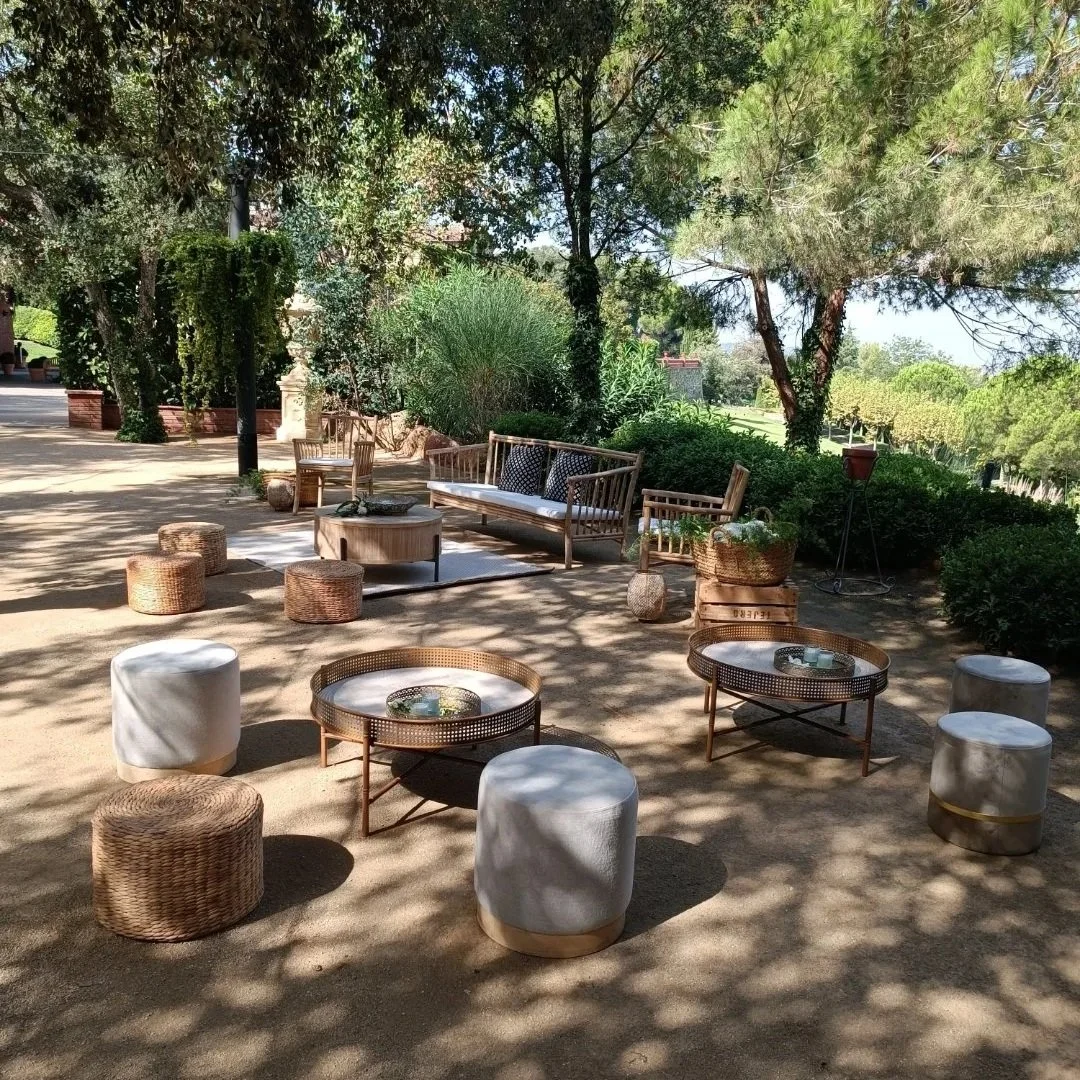 Área de descanso al aire libre con sillas, mesas y cojines de mimbre en sombra de árboles.