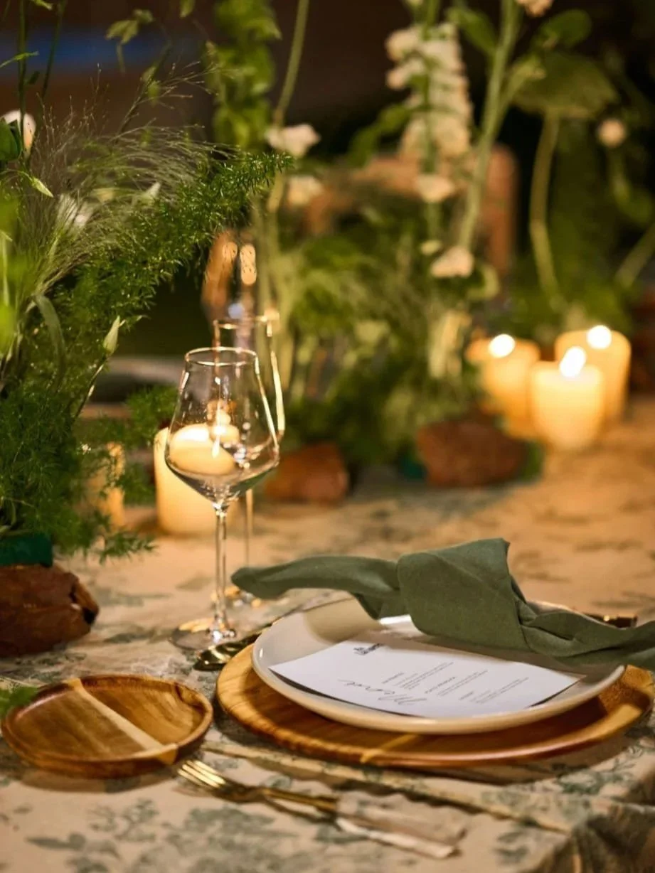Mesa decorada con platos, cubiertos, una copa de vino, un menú, un mantel con un diseño floral, y velas en un ambiente con muchas plantas y flores tropicales.