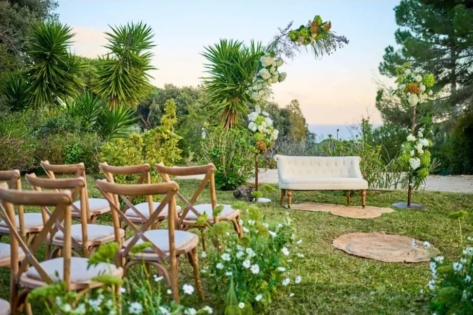 Una ceremonia al aire libre con sillas de madera, un sofá de estilo vintage y decoraciones florales en un entorno natural, posiblemente en la playa o cerca del mar.