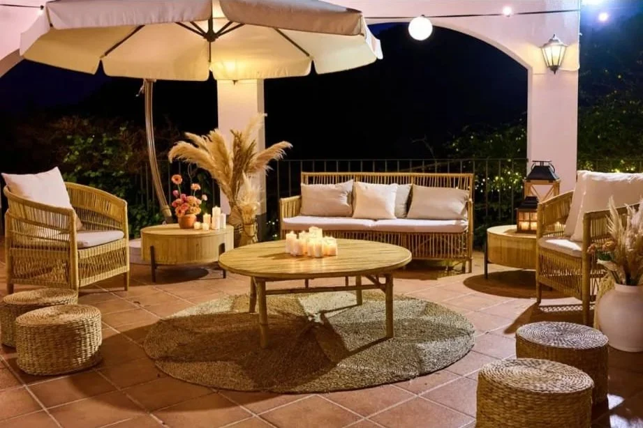 Espacio de terraza decorado con mobiliario de mimbre, cojines blancos, lámparas, velas y plantas en jarrones, ambientado para la noche.