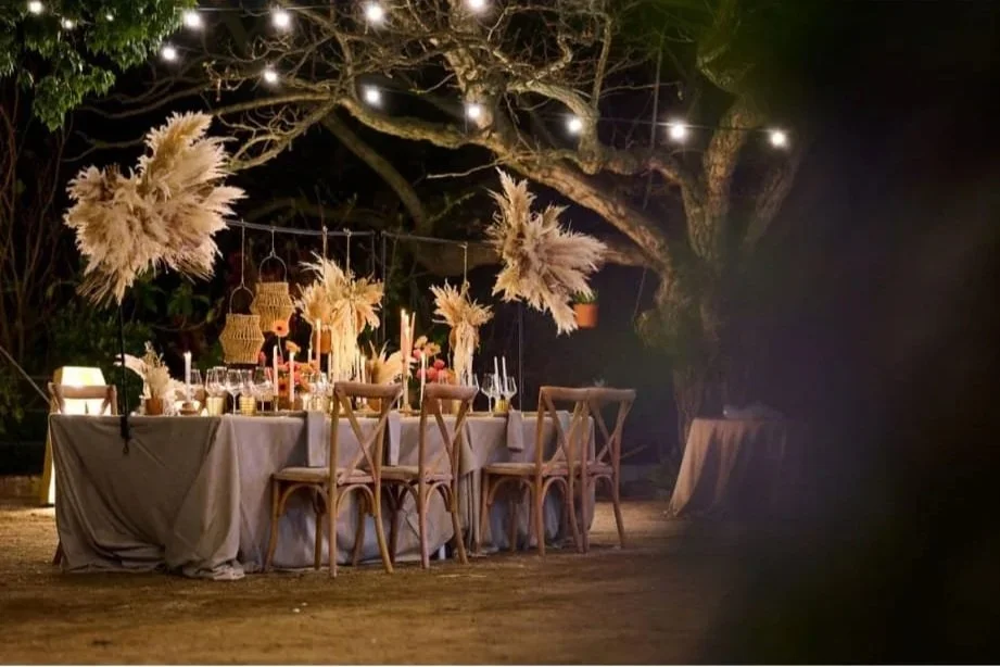 Mesa decorada para una celebración nocturna con guirnaldas de luces, adornos de flores y velas en un entorno al aire libre con árboles y ambiente cálido.