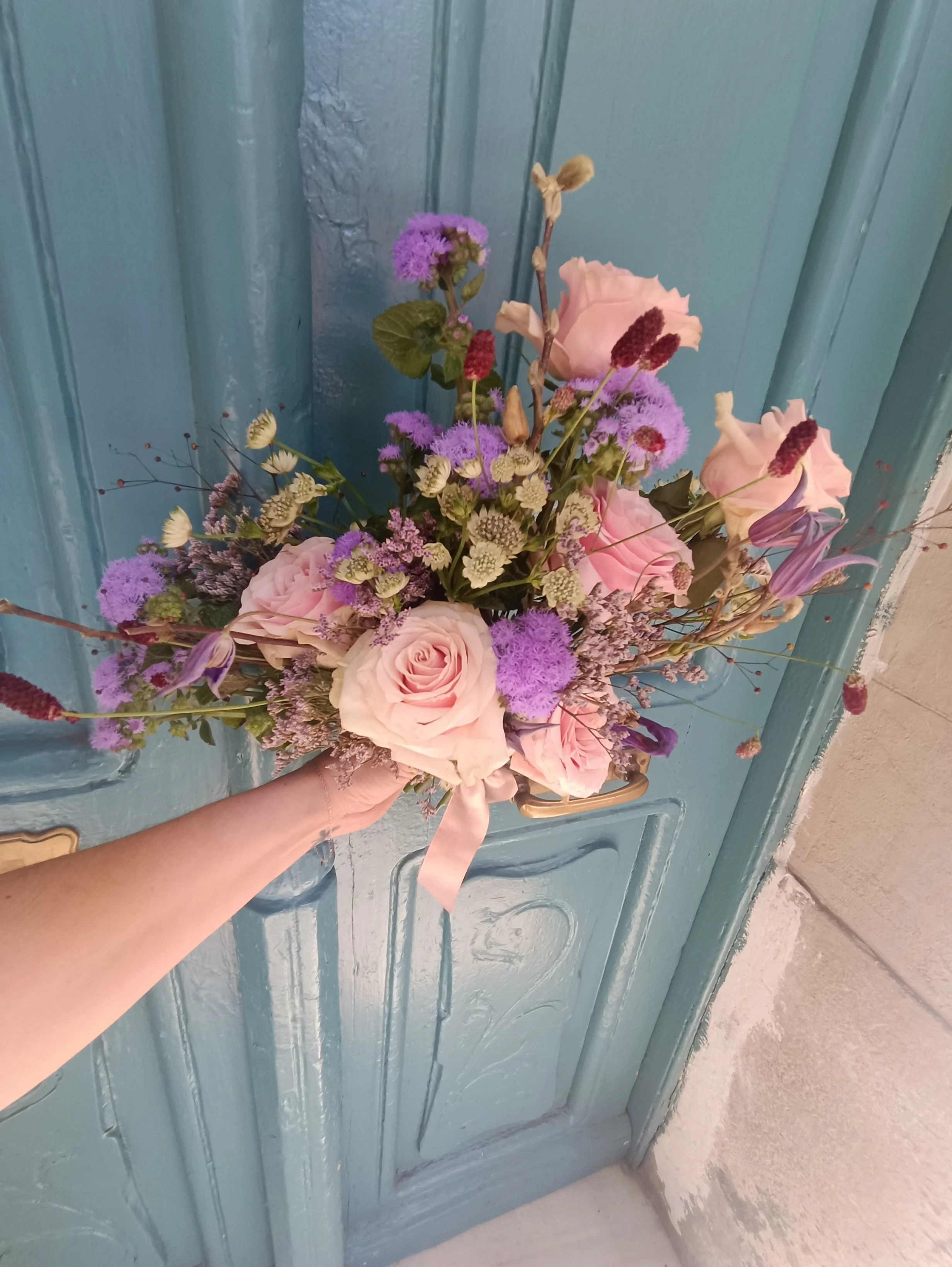 Mano sosteniendo un ramo de flores frescas, incluyendo rosas rosadas, lavanda y otros flores en tonos morados y verdes, frente a una puerta azul