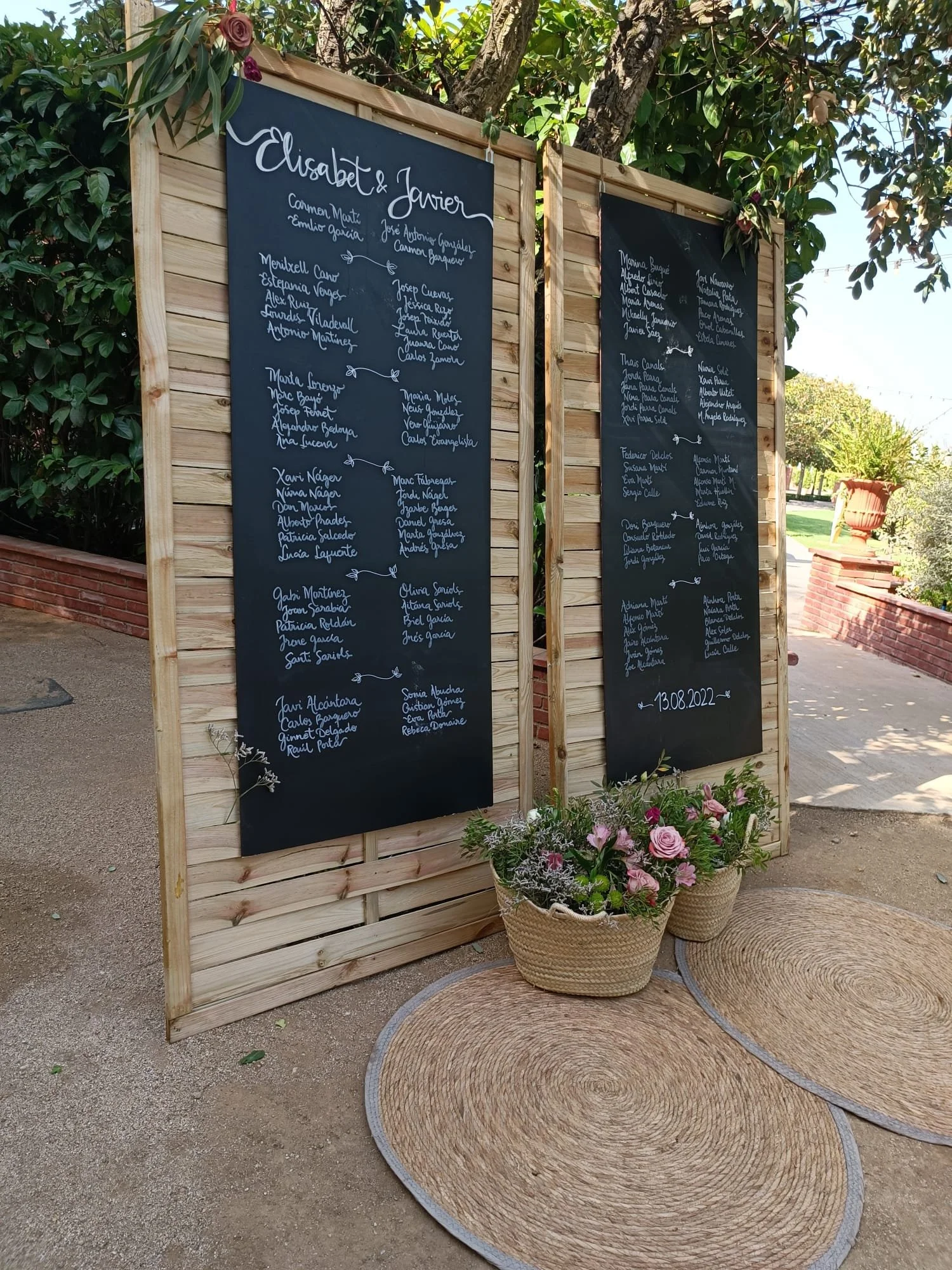 Carteles nupciales escritos a mano con nombres y fecha, decorados con flores rosas y verdes, enmarcados en madera, situados en un ambiente al aire libre con plantas y árboles.