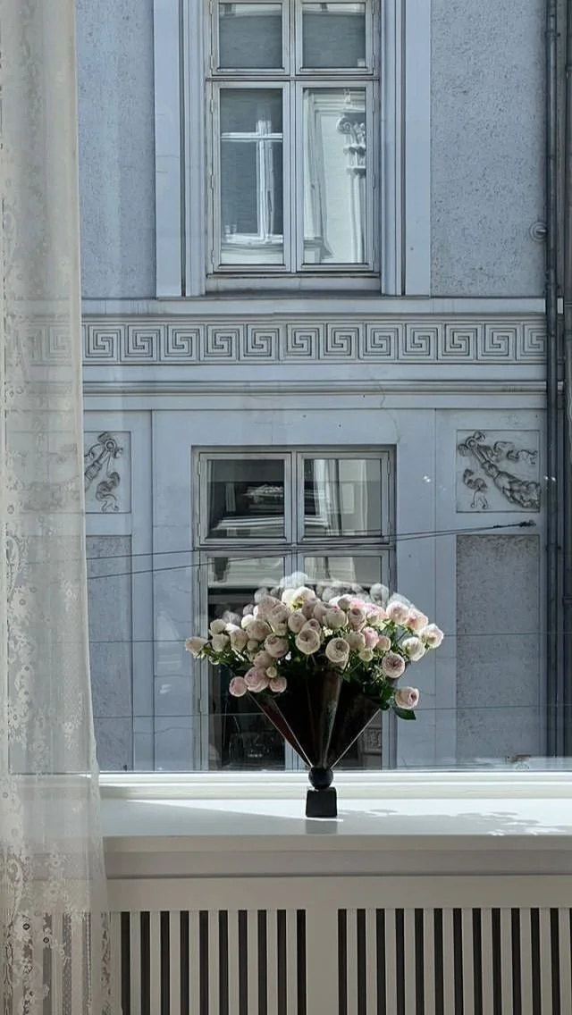 Florero con flores blancas en una ventana con vista a un edificio con ventanas y detalles decorativos.