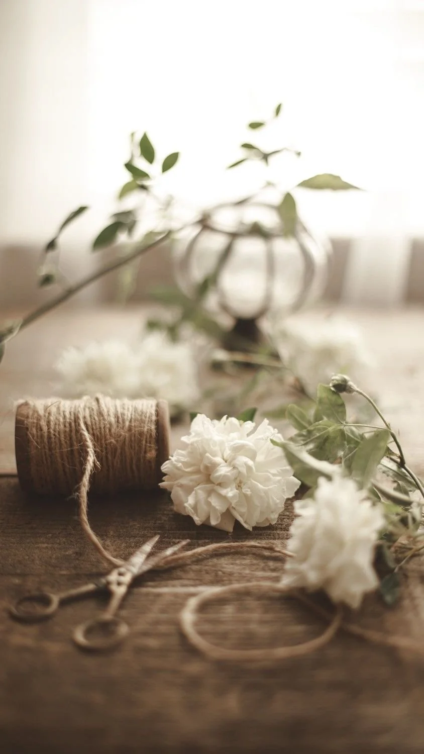 Material de manualidades con cuerda, tijeras, flores blancas y follaje en una mesa de madera, con un fondo difuso.