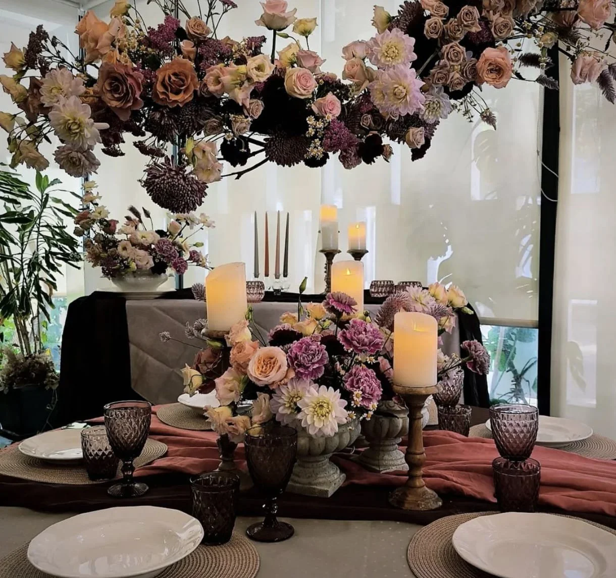 Decoración elegante con flores en tonos pastel, velas y cristalería en una mesa preparada para evento.