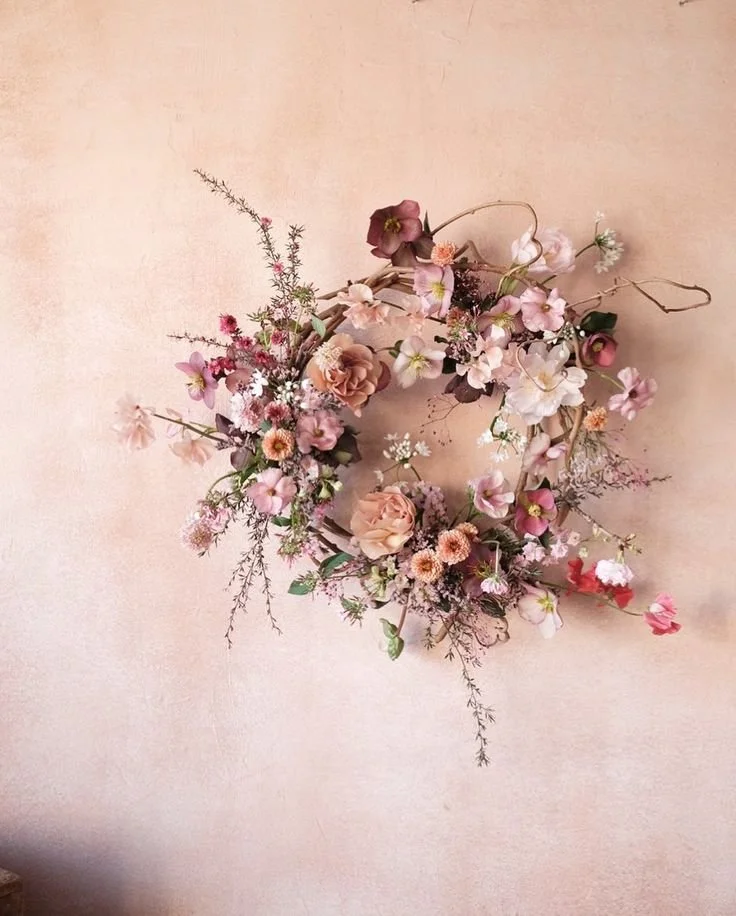 Corona de flores en tonos rosa, beige y blanco colgada en una pared de color crema.