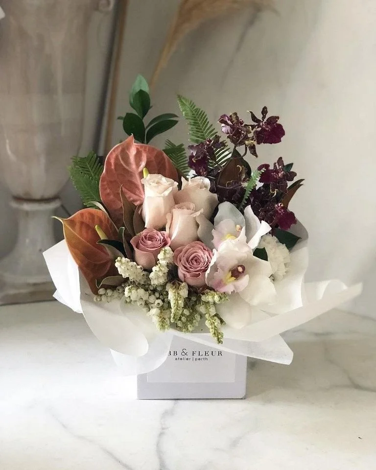 Arreglo de flores con rosas rosadas, orquídeas blancas, hojas verdes, y plantas moradas, en caja blanca con la inscripción 'B & Fleur atelier Perth'.