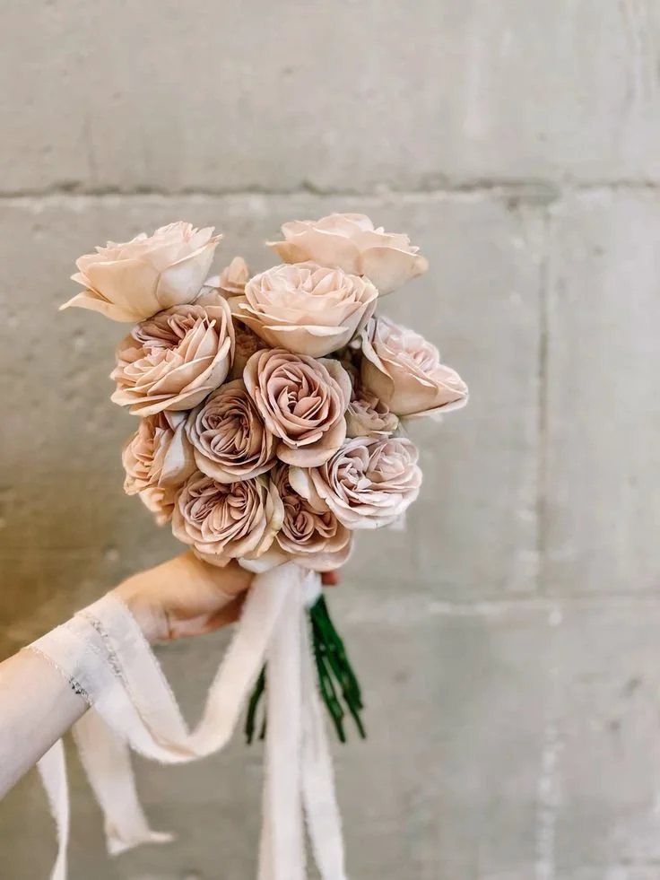 Ramo de rosas de color rosa pálido sostenido con una mano frente a una pared de concreto.