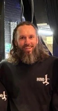 A man with long hair and a beard smiling, wearing a black shirt with a logo on it, in an indoor setting with dark curtains and some lights in the background.
