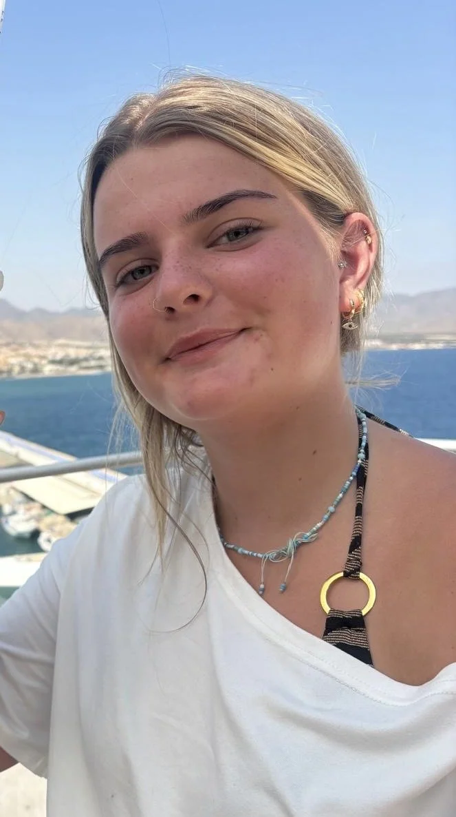 Close-up of a young woman with blonde hair smiling outdoors, with water and distant mountains in the background.