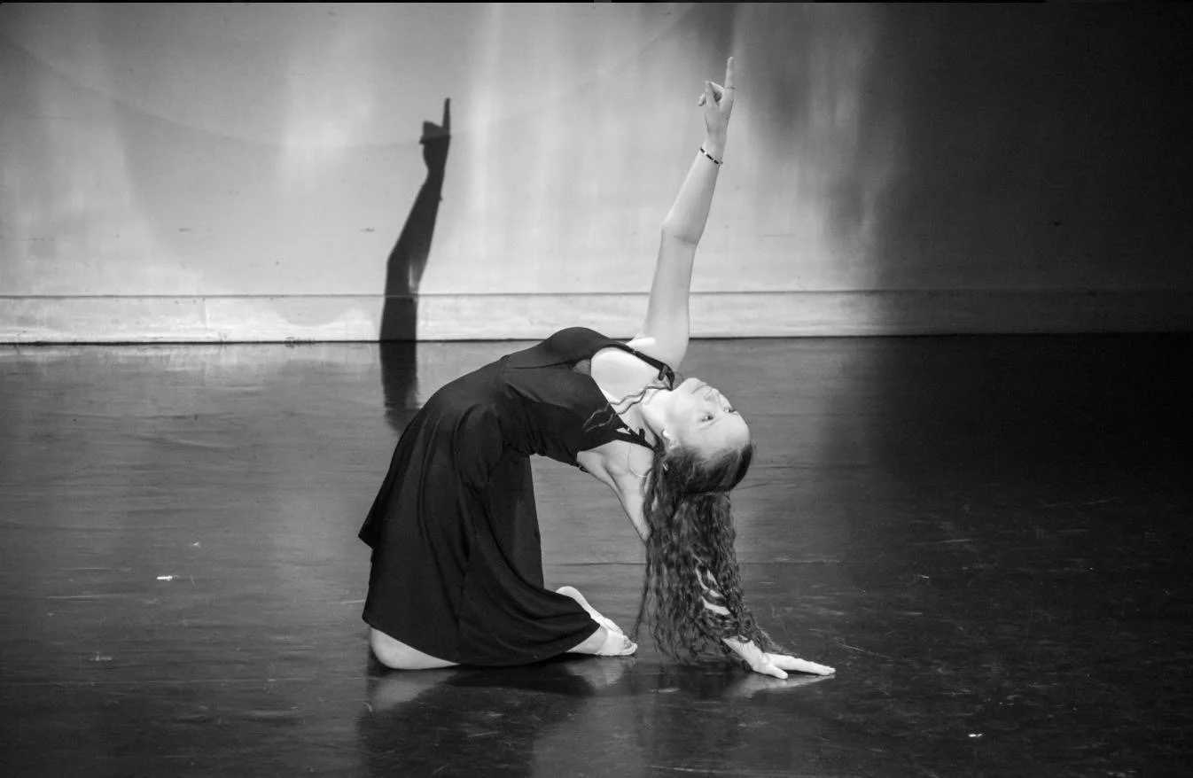 A woman with long curly hair performing a dance move on a stage, kneeling on one knee with her head tilted back, one arm extended upward and the other arm resting on the floor.