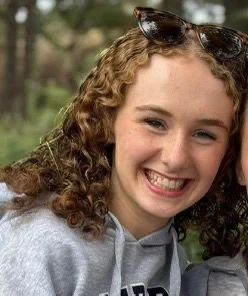 A dance performance on stage with four girls in colorful costumes, surrounded by an audience of young children and a group of people in black shirts in the background. The backdrop displays the logo 'KBD Dance.'