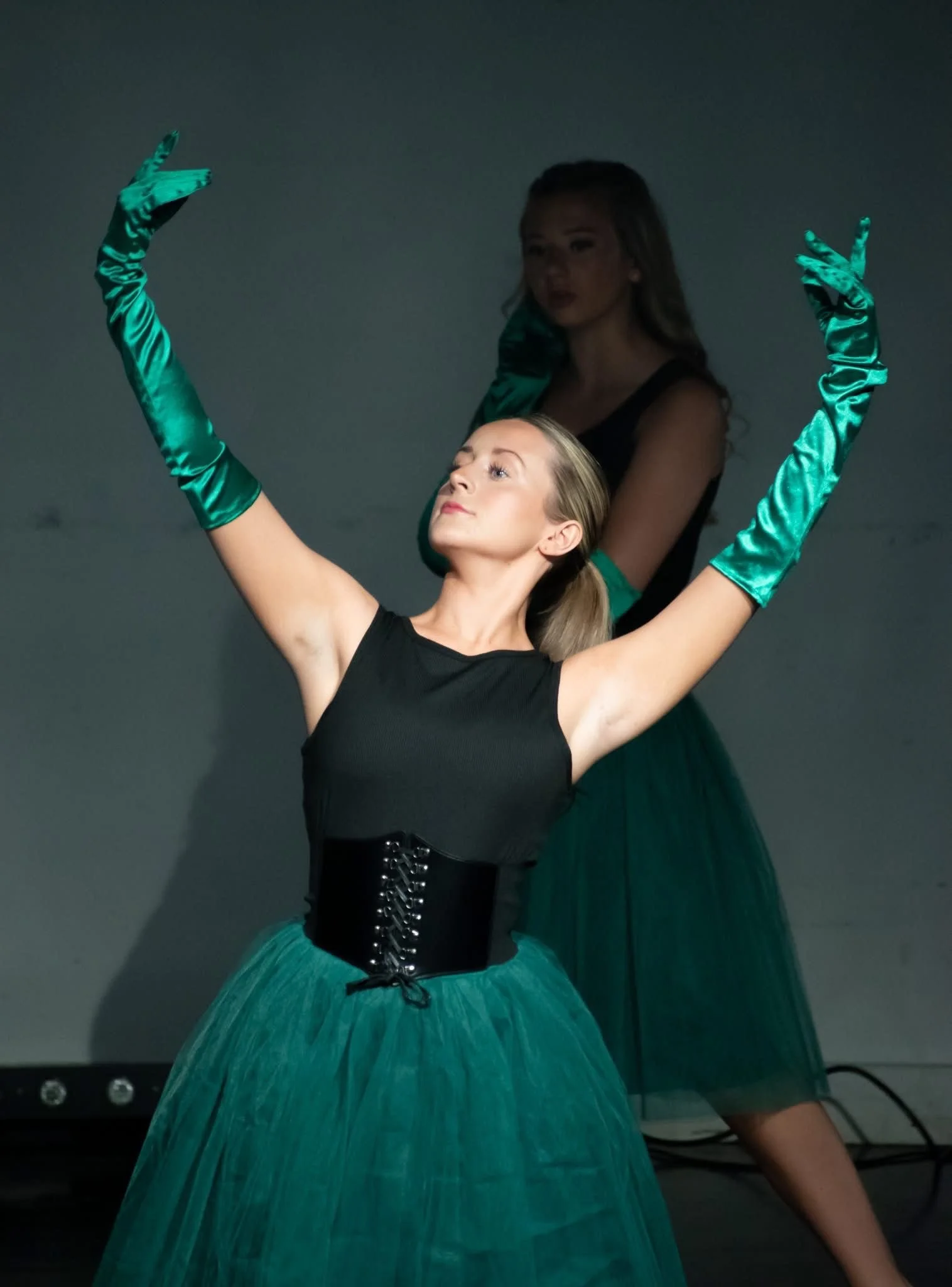 Two women in ballet costumes, with one in the foreground wearing a black top, corset, and teal tutu, and the other in the background wearing a black top, teal gloves, and a teal tutu, performing a dance pose against a plain background.