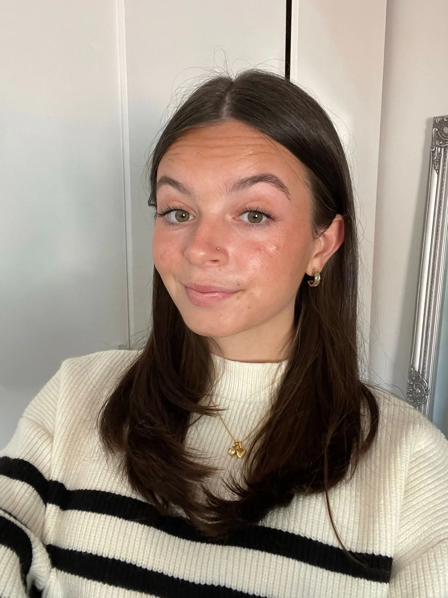 A young woman with medium-length dark brown hair, green eyes, and a nose piercing, smiling while taking a selfie in a room with a mirror and a white wall.