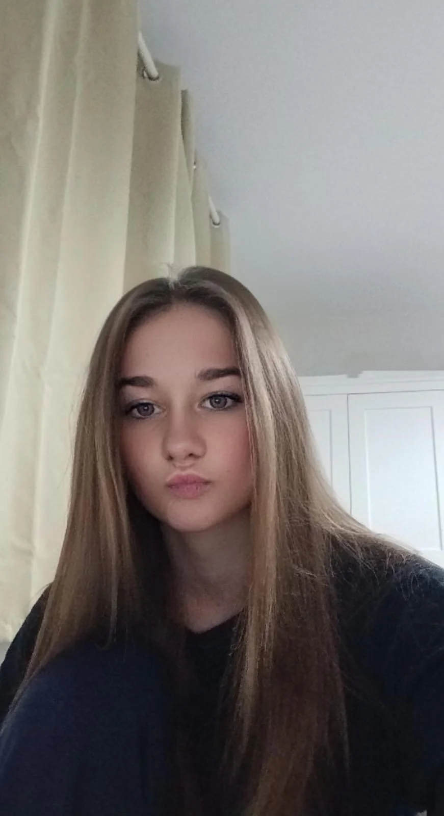 A young woman with long, light brown hair and light-colored eyes smiling softly indoors, with cream curtains and white furniture in the background.