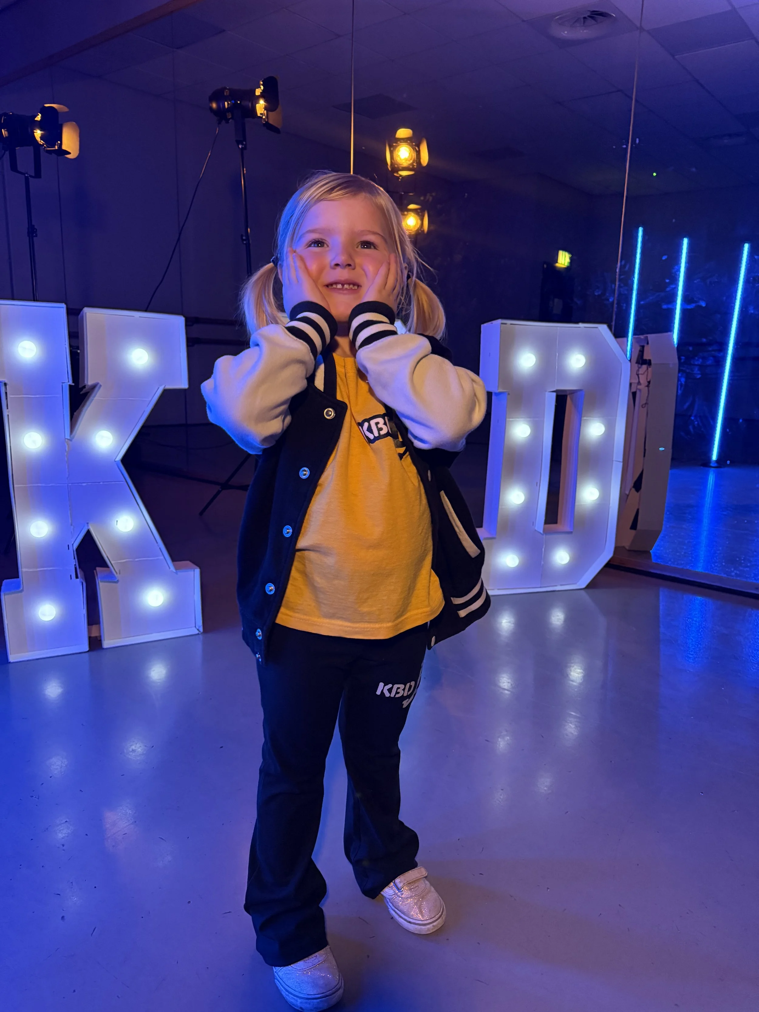 A young girl with blonde hair in pigtails standing in front of illuminated marquee letters spelling 'KID' in a dark room, with her hands on her face and smiling.