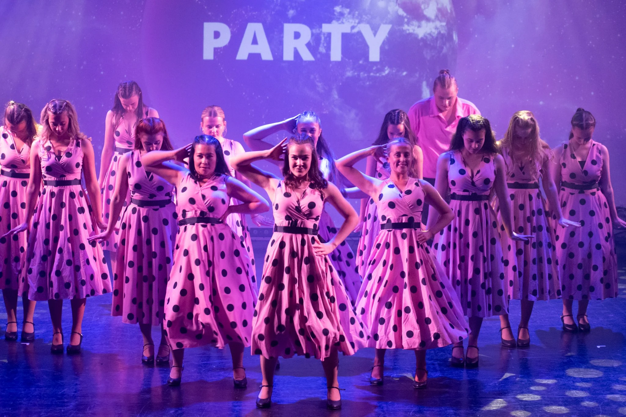 Group of women and girls in matching pink polka dot dresses performing a dance on stage with a purple background that says "PARTY."