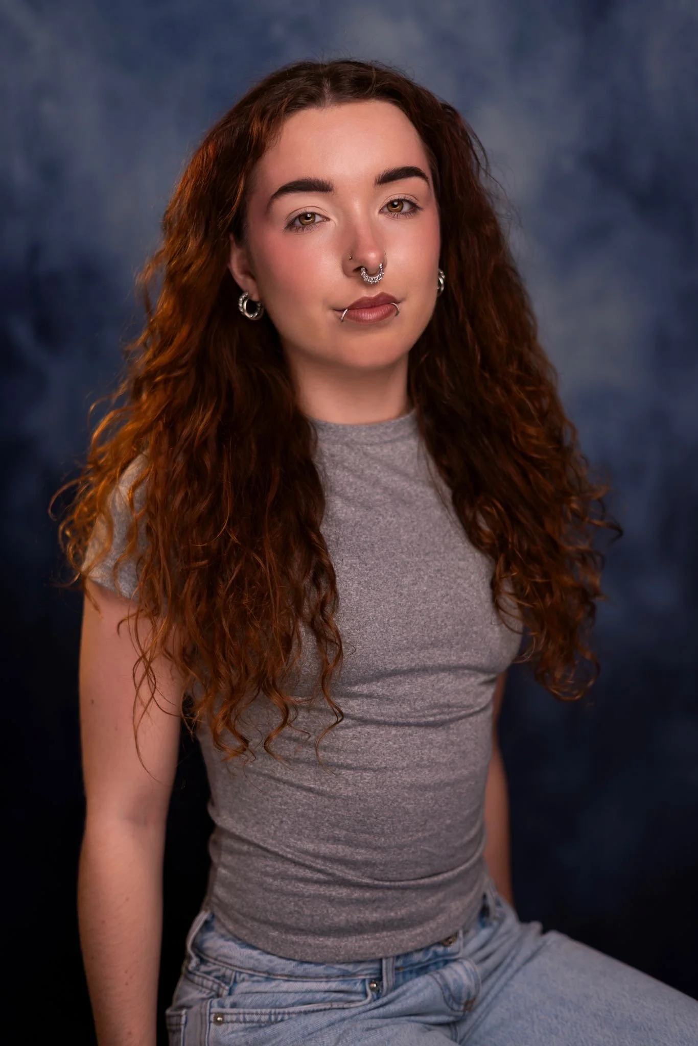 A young woman with long curly red hair and multiple facial piercings, wearing a gray t-shirt and jeans, against a dark blue background.