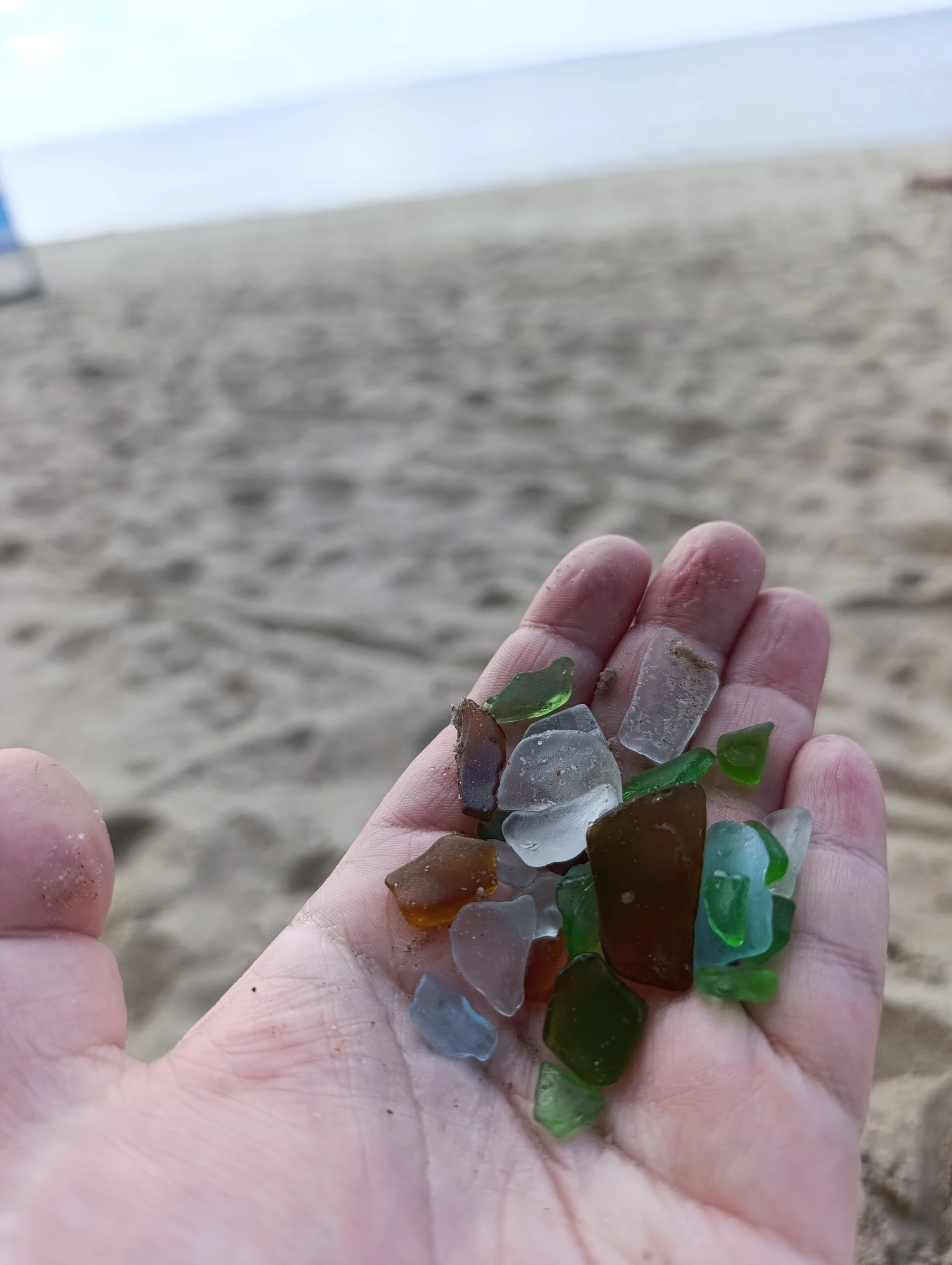 A hand holding colorful sea glass pieces on a sandy beach with the ocean in the background.