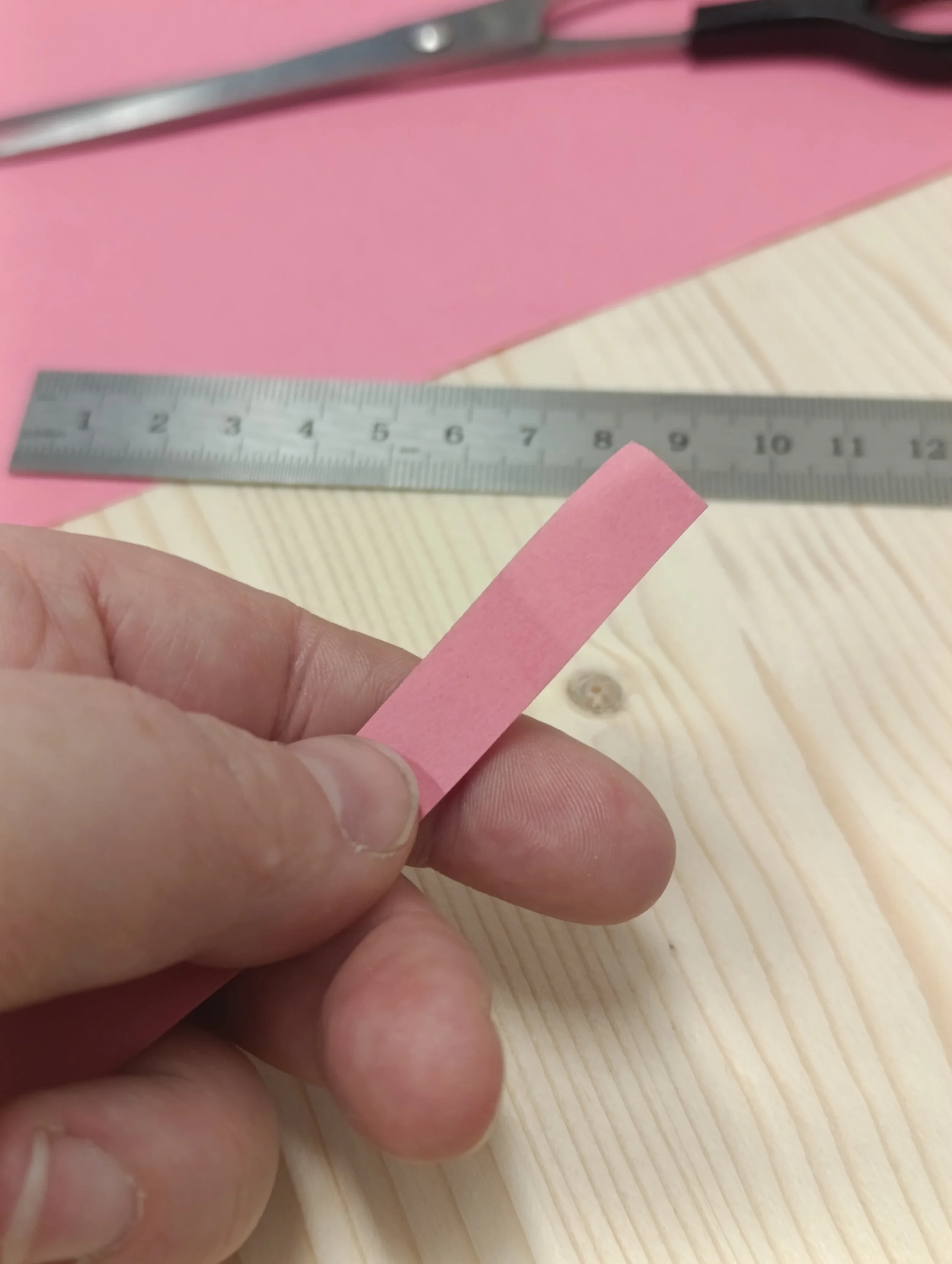 Close-up of a person's hand holding a small piece of pink paper near a metal ruler on a light wooden surface.