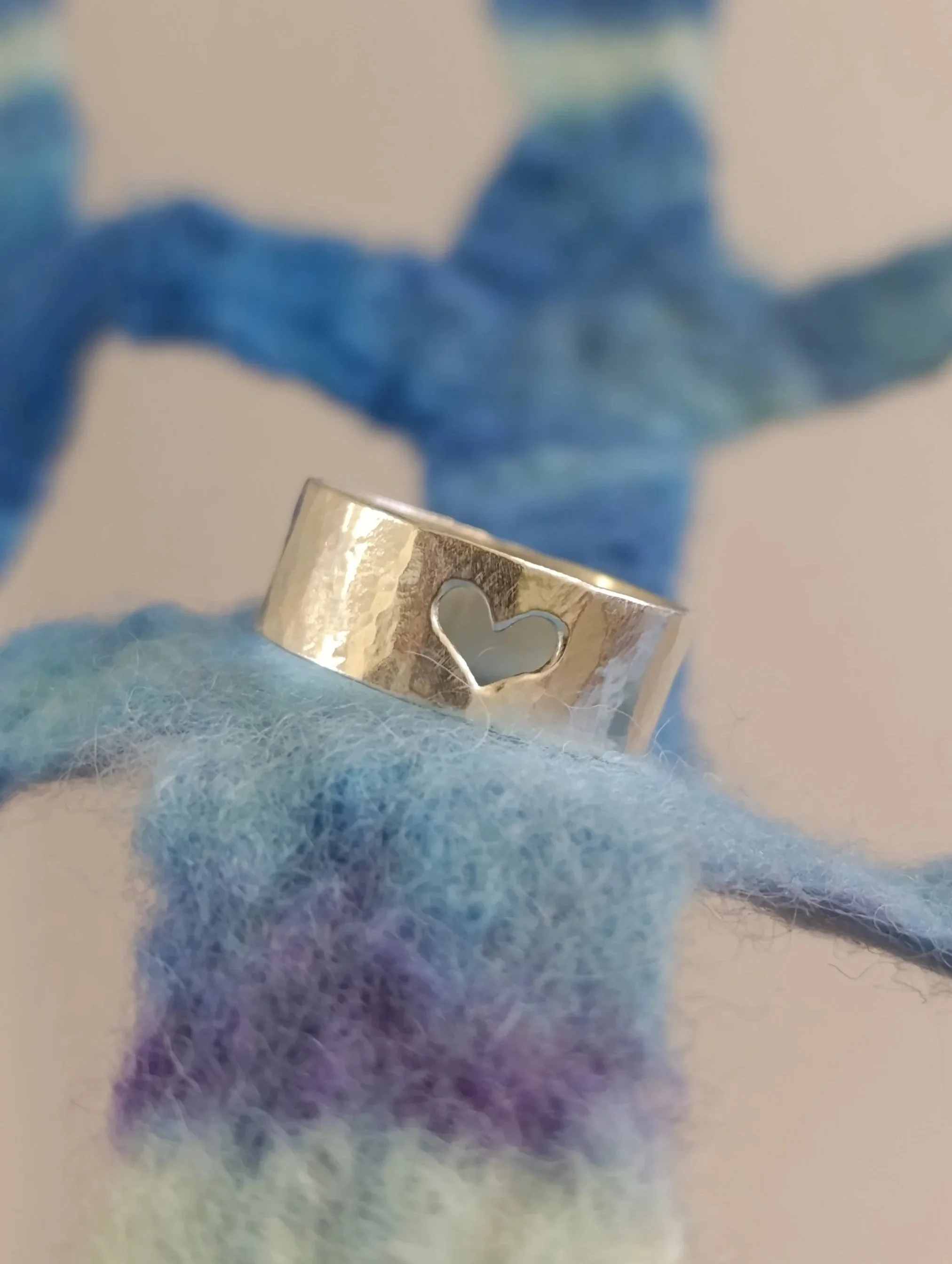 A silver ring with a heart-shaped cutout, resting on soft, multicolored wool yarn with a small blue and lavender teddy bear in the background.