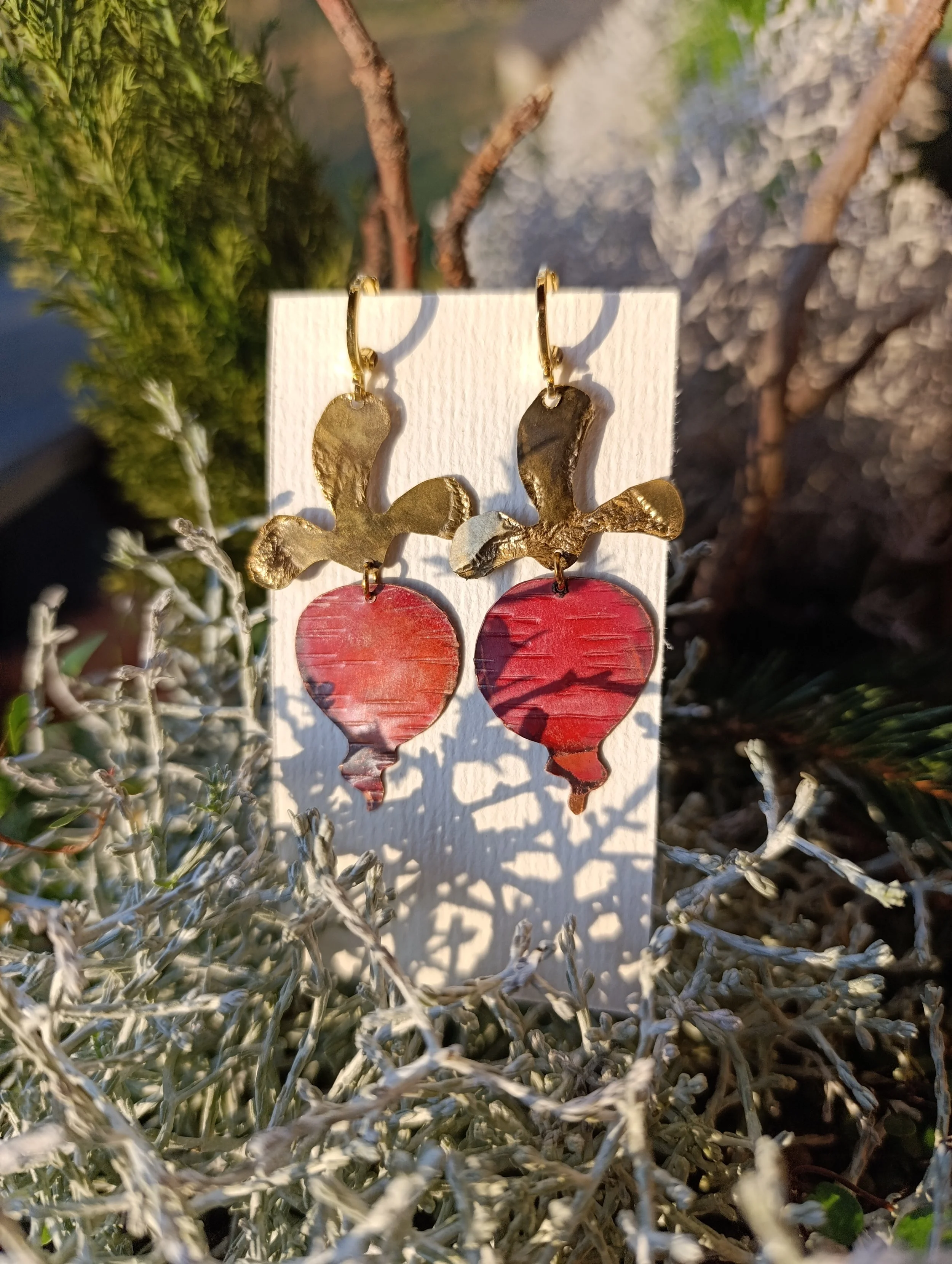 Gold and red strawberry-shaped earrings displayed on a white card, surrounded by greenery and dried plants.