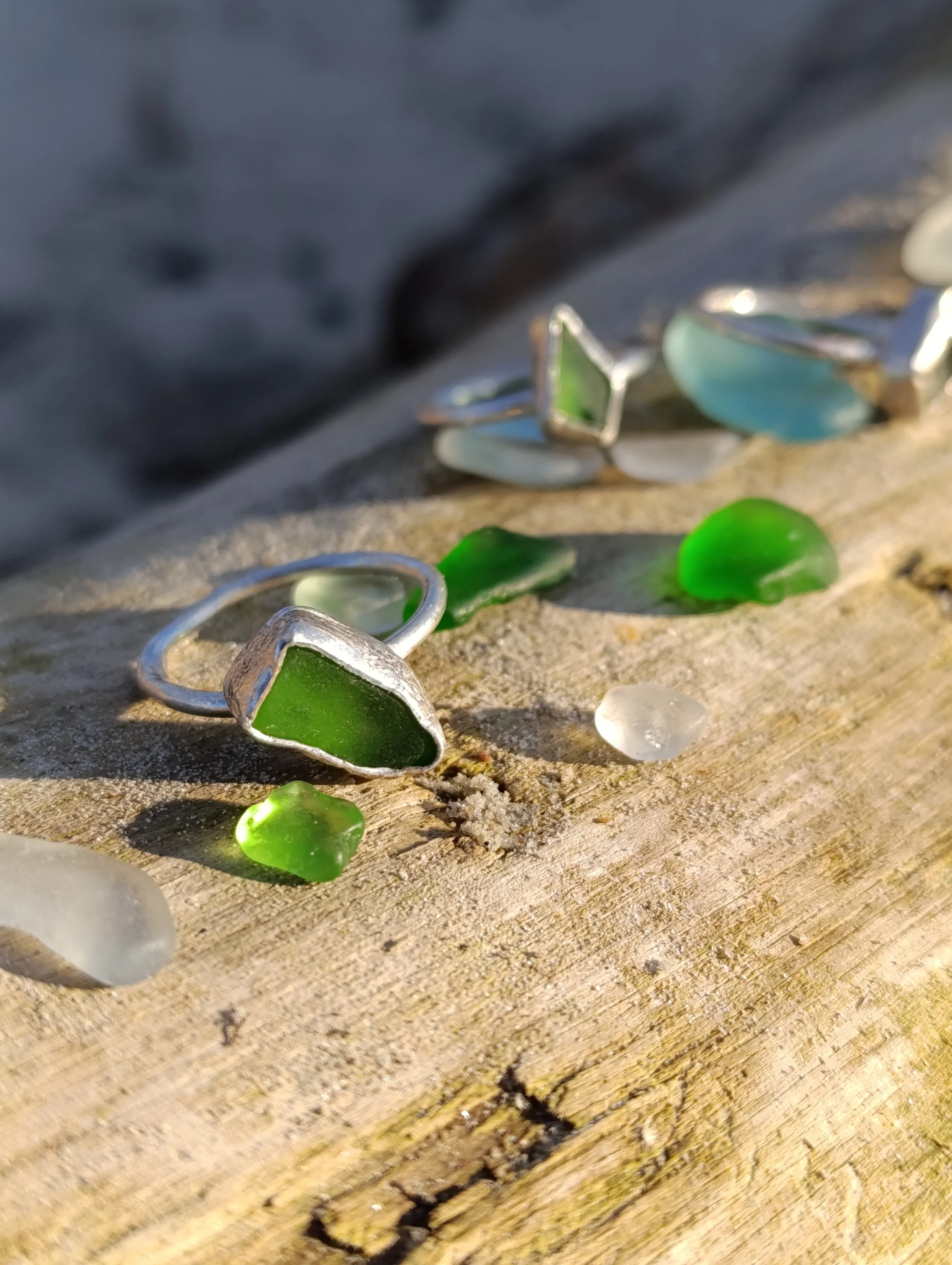 Colorful sea glass and silver rings arranged on a sandy wooden surface with sunlight casting shadows.