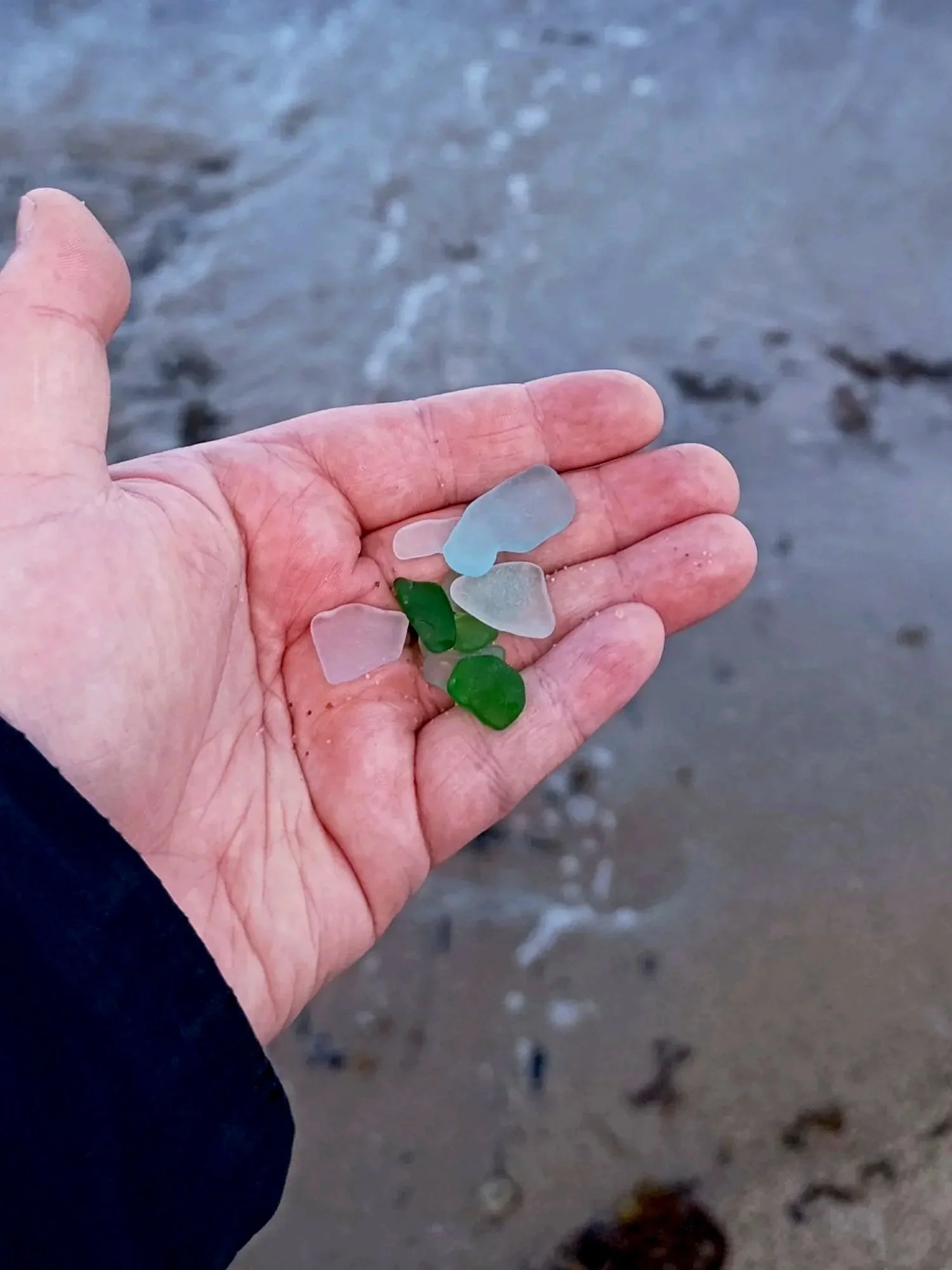 A person's hand holding several pieces of colorful sea glass near the shoreline with water and sand in the background.