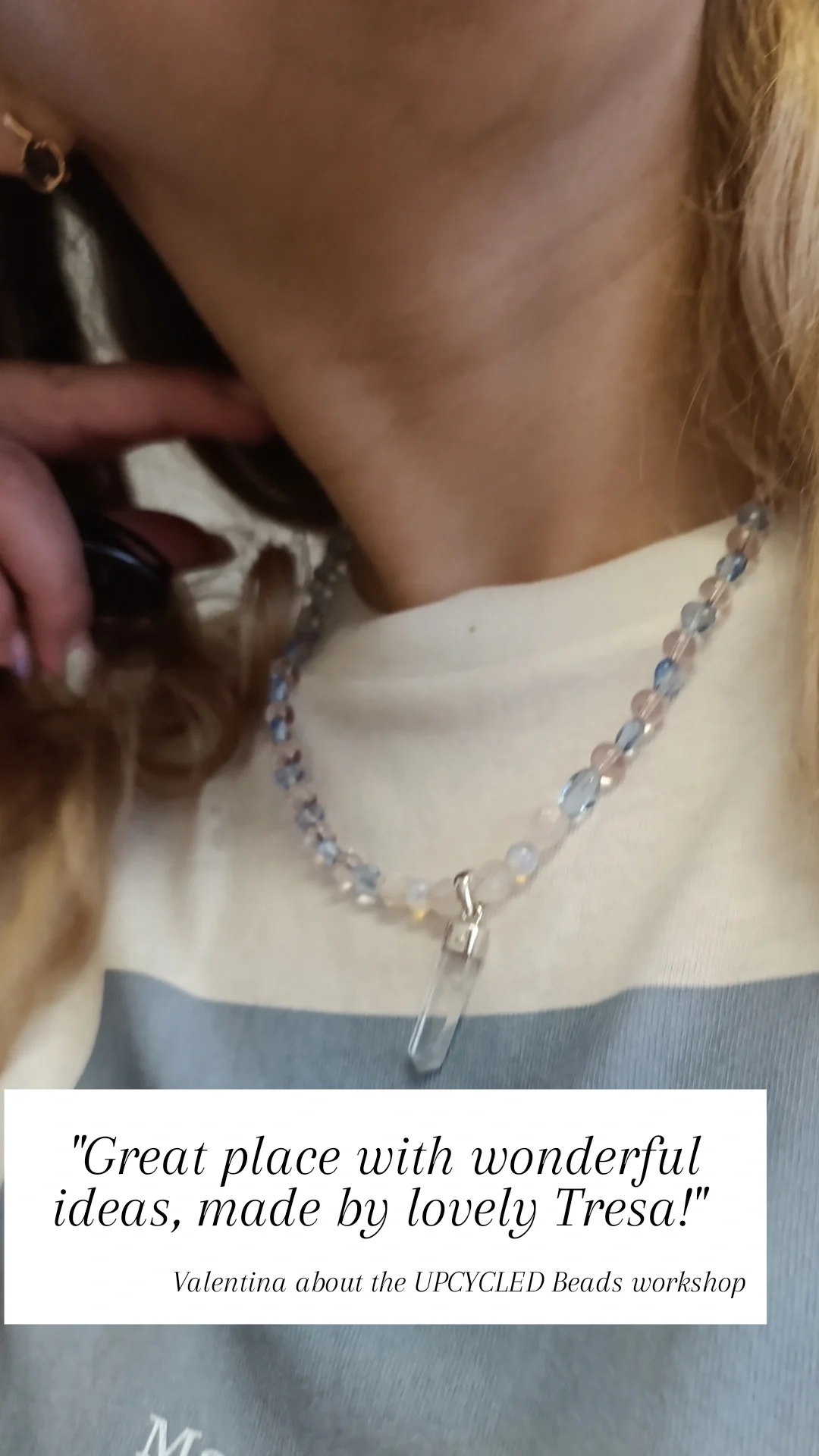 Close-up of a person's neck and upper chest wearing a beaded necklace with a clear pendant. The person has a light-colored shirt with partially visible text and blonde hair. A caption overlay reads: "Great place with wonderful ideas, made by lovely Tresa!" Valentina about the UPCYCLED Beads workshop.