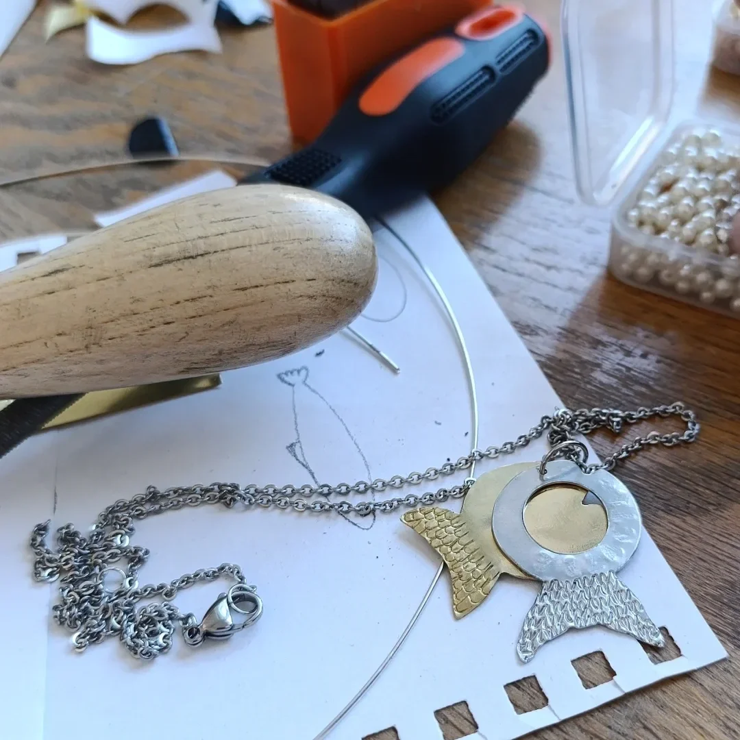 Jewelry-making supplies on a wooden table, including a chain, metal charms of a fish, moon, and an angel, a wooden handle tool, a craft wire, a small screwdriver, and a container of pearl beads.