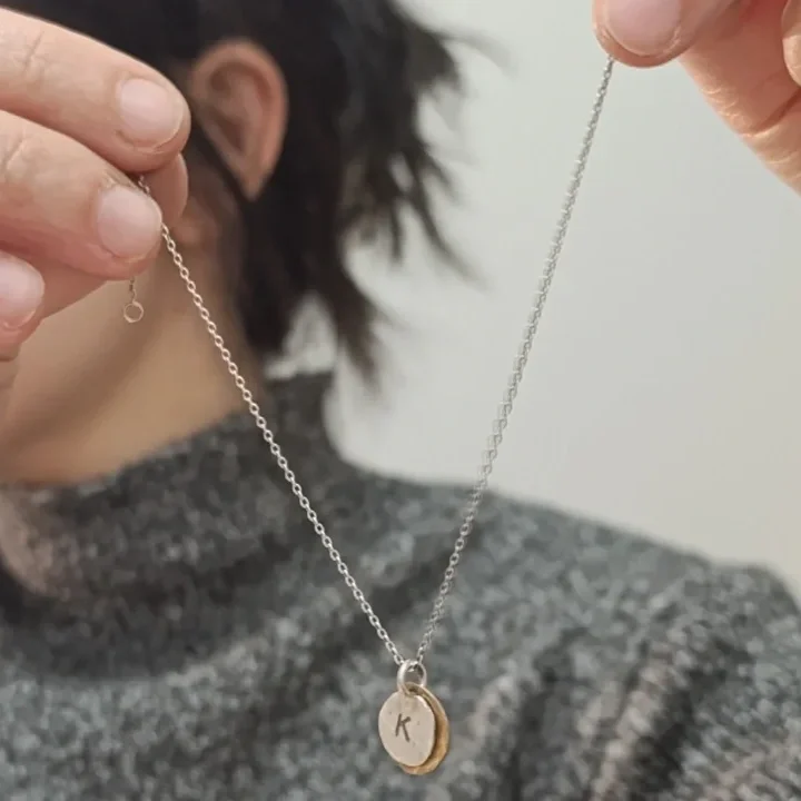 Person holding a silver chain necklace with a small oval pendant engraved with the letter 'K'.