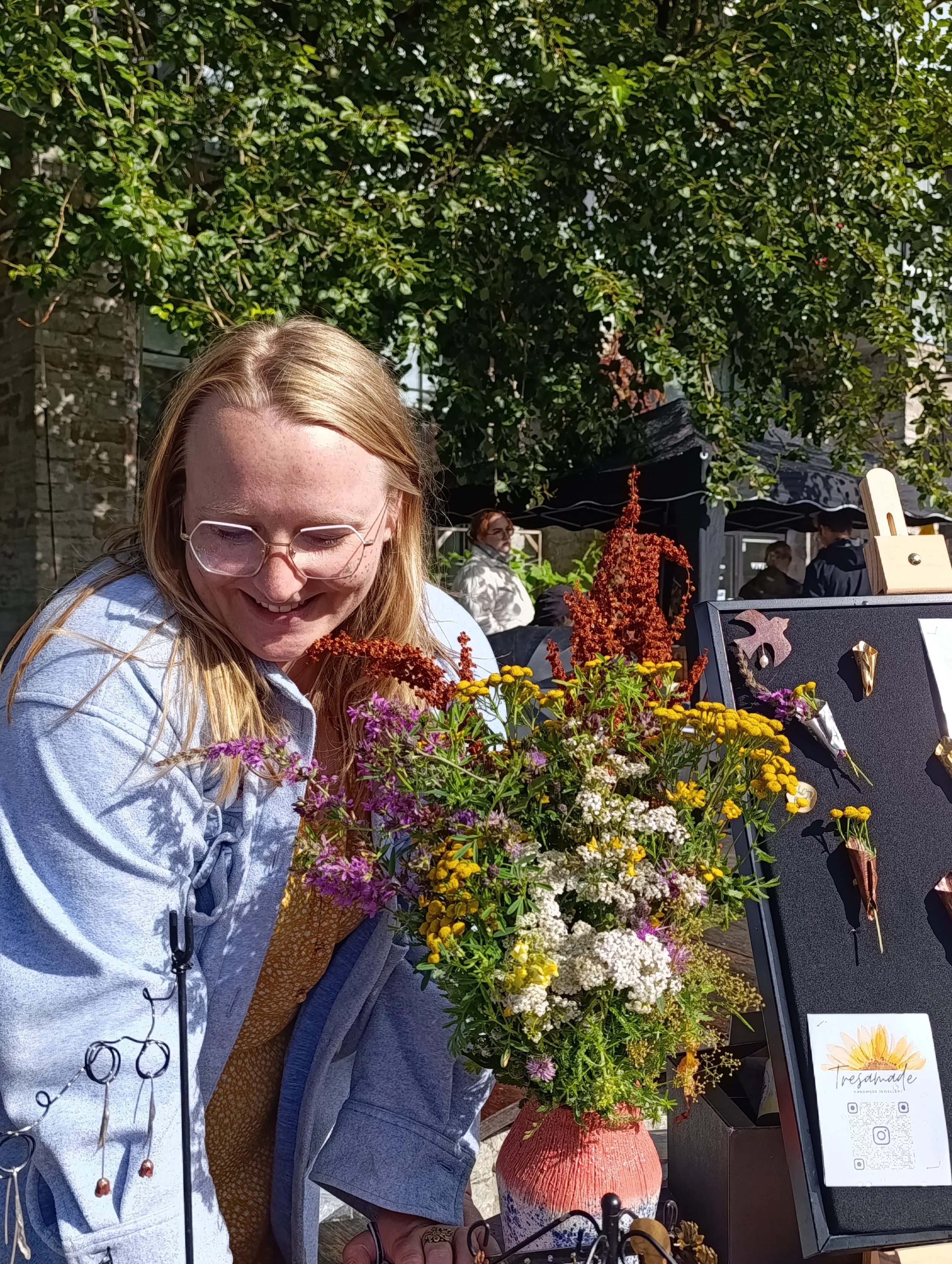 A woman with glasses smiling at a flower display at an outdoor market or craft fair. She is standing next to a colorful bouquet of wildflowers in a red vase, with various art and jewelry on display nearby.
