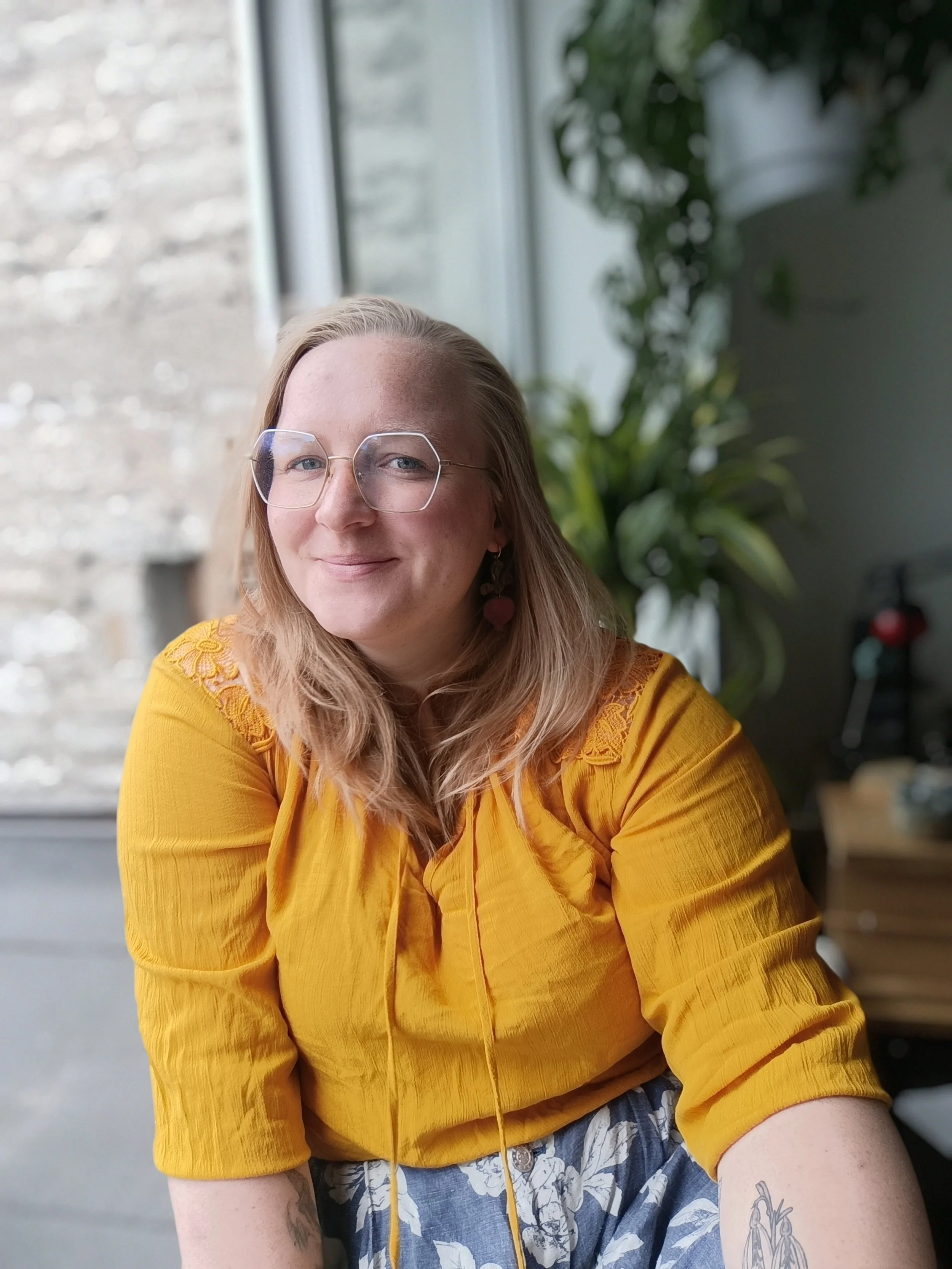 A woman with blonde hair, wearing glasses and a yellow top, sitting indoors near a window with a large green plant behind her.