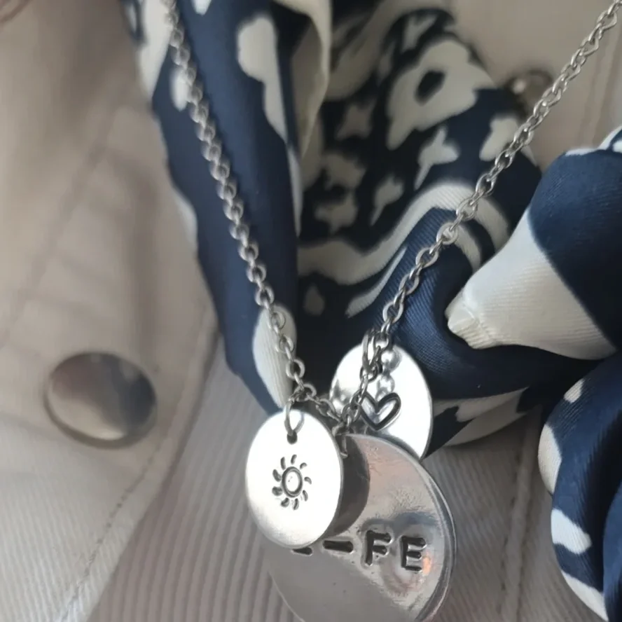 Close-up of a silver necklace with multiple charms, including a sun, a heart, and a round tag with the letters "E-FE" engraved, resting on a black and white patterned fabric.