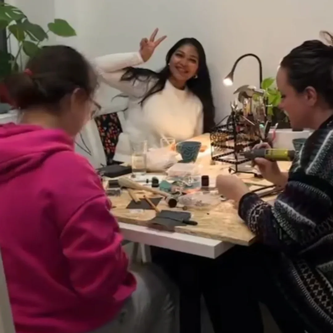 Group of young women sitting around a table, engaging in a craft or makeup activity, with one woman smiling and making a peace sign.