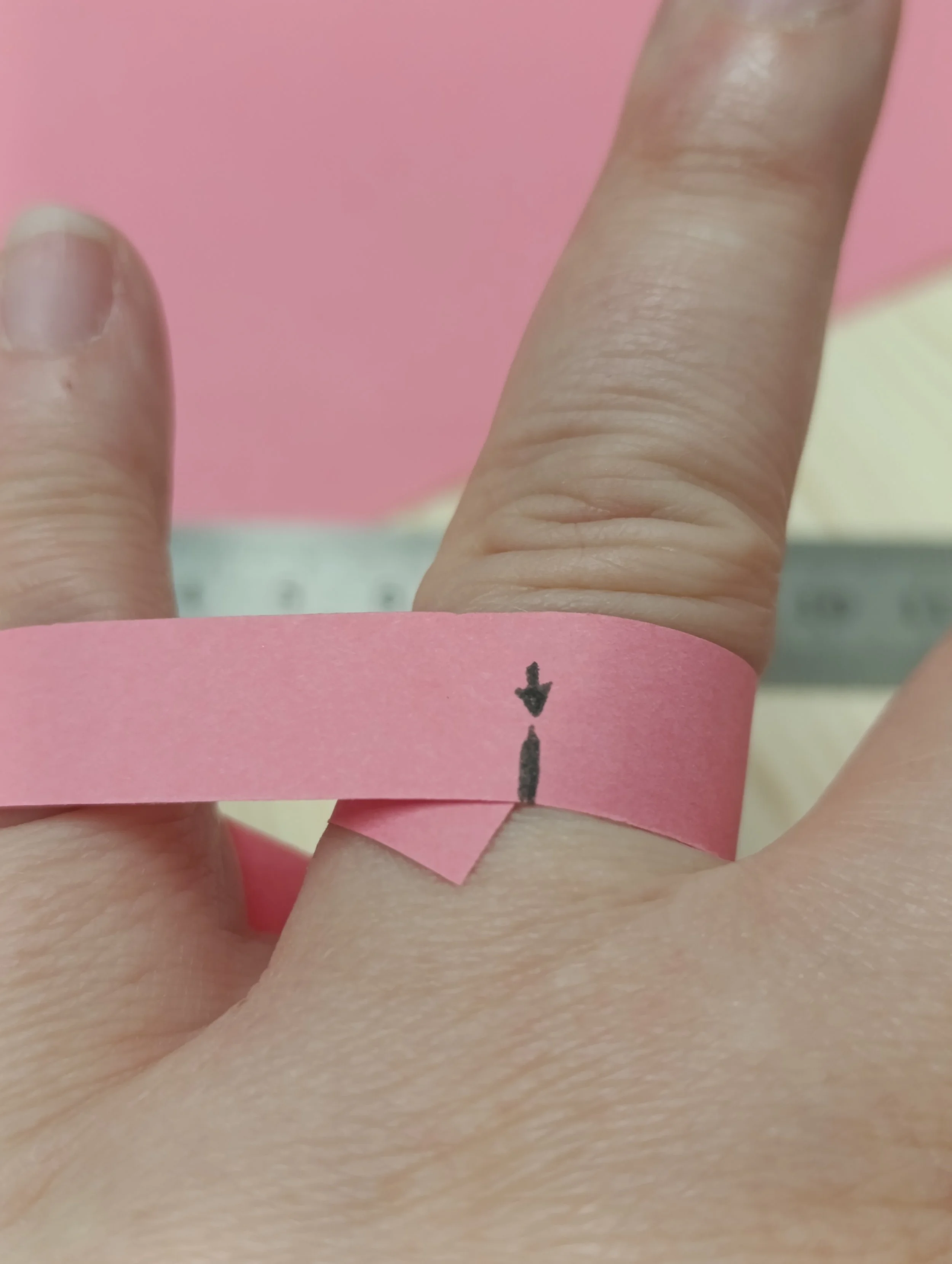 A person's hand with a pink paper ring around the ring finger, with a small black arrow pointing towards the finger.