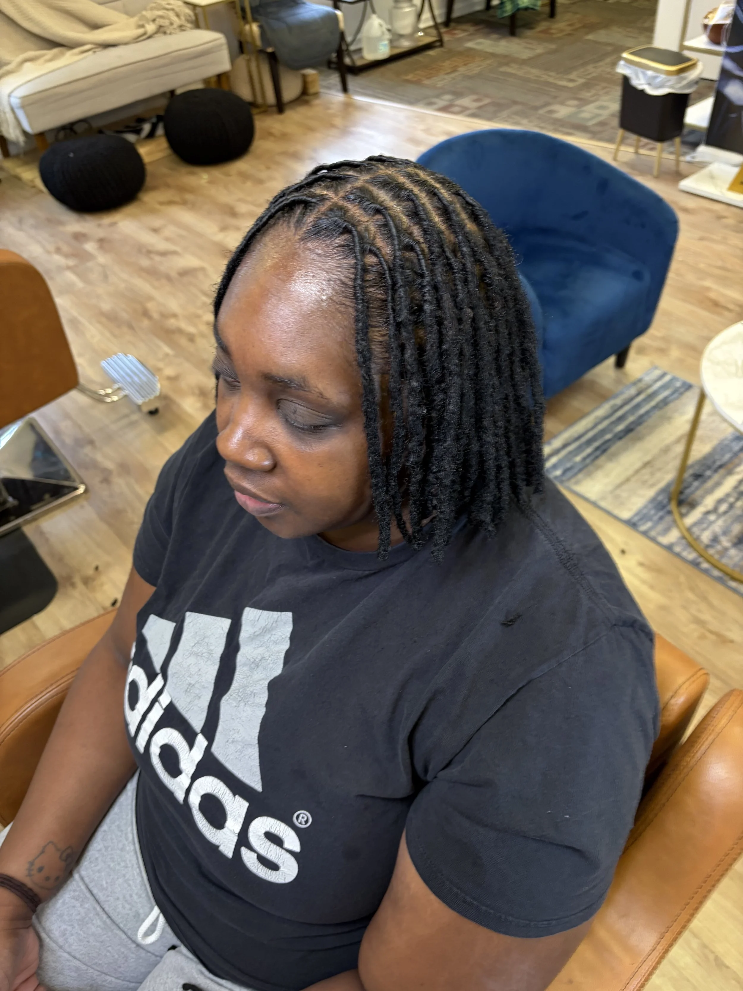 A woman with styled dreadlocks decorated with colorful beads, sitting with her arms crossed, wearing a gray T-shirt, and a smartwatch on her left wrist, in a salon or similar setting.