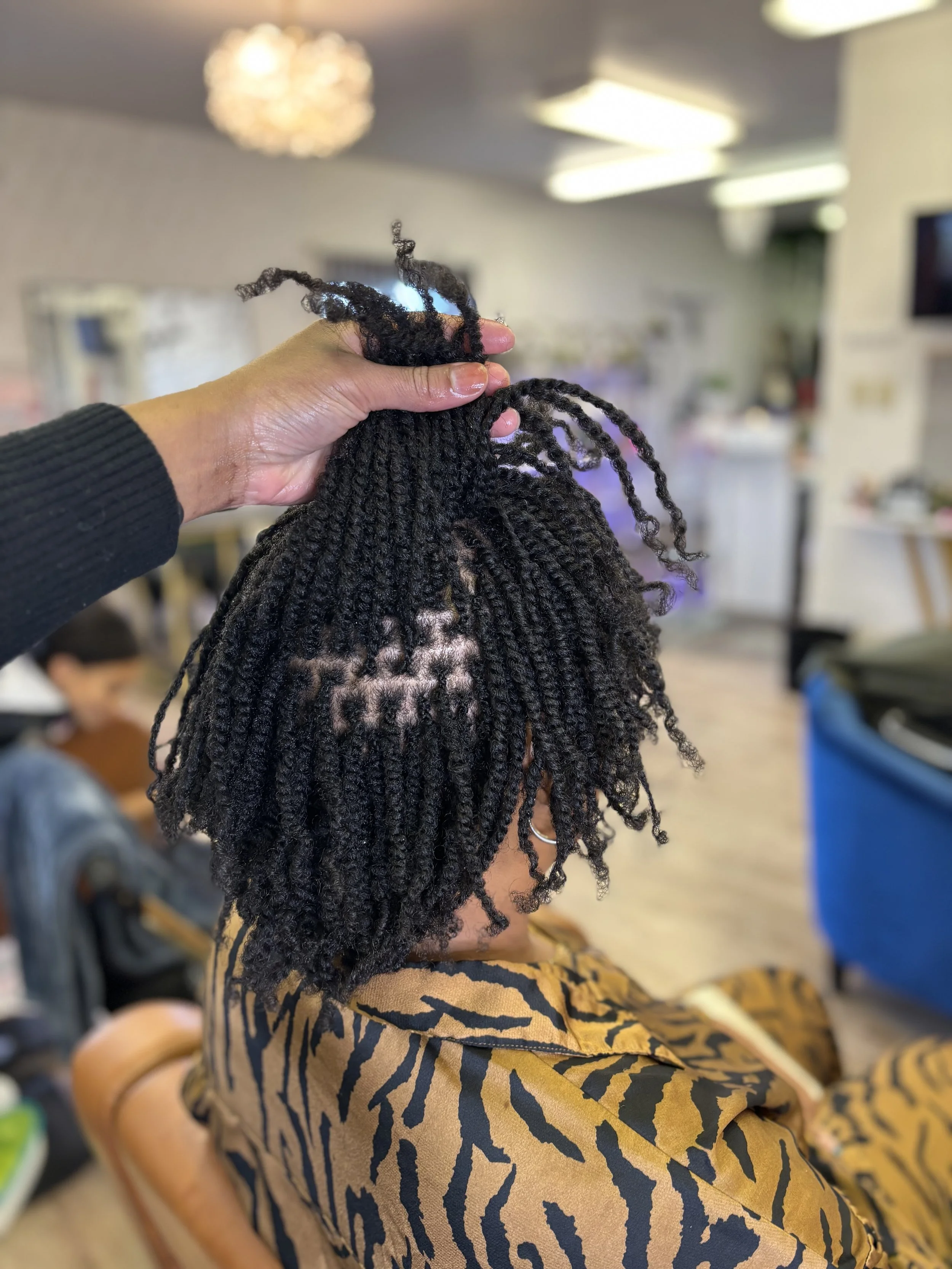 Person holding a section of freshly styled twisted dreadlocks above a woman with shoulder-length dreadlocks seated in a salon.