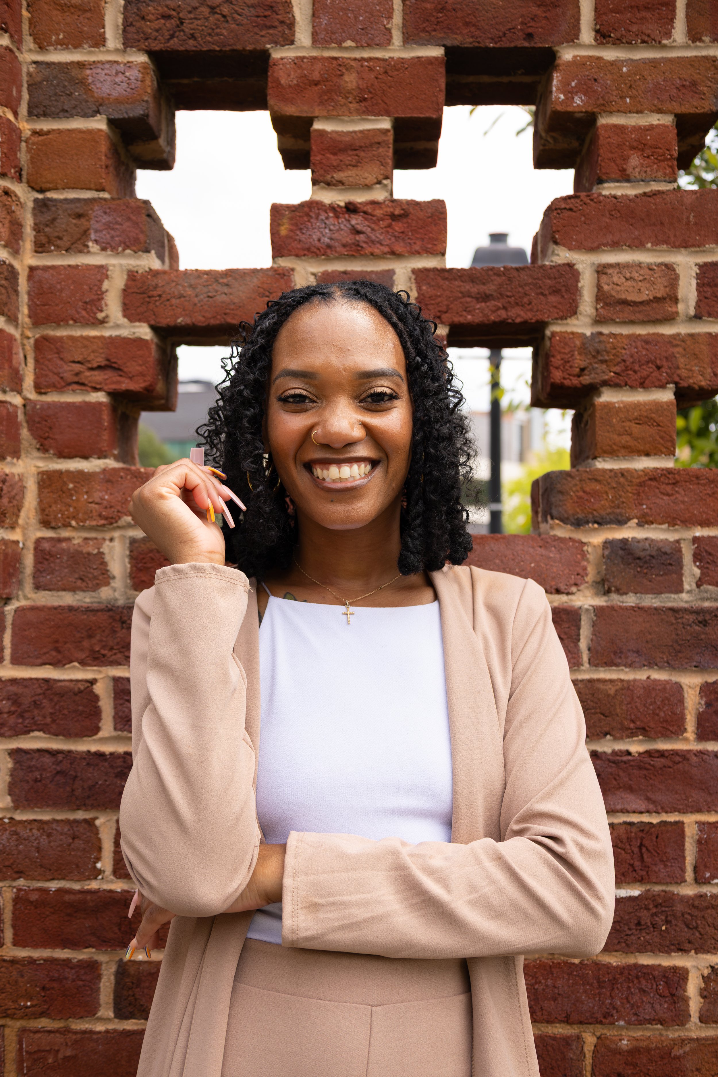 A smiling woman with short curly hair, hoop earrings, a nose ring, a necklace with an ankh, a black top, a blue blazer, and black pants, standing outdoors with green trees in the background.