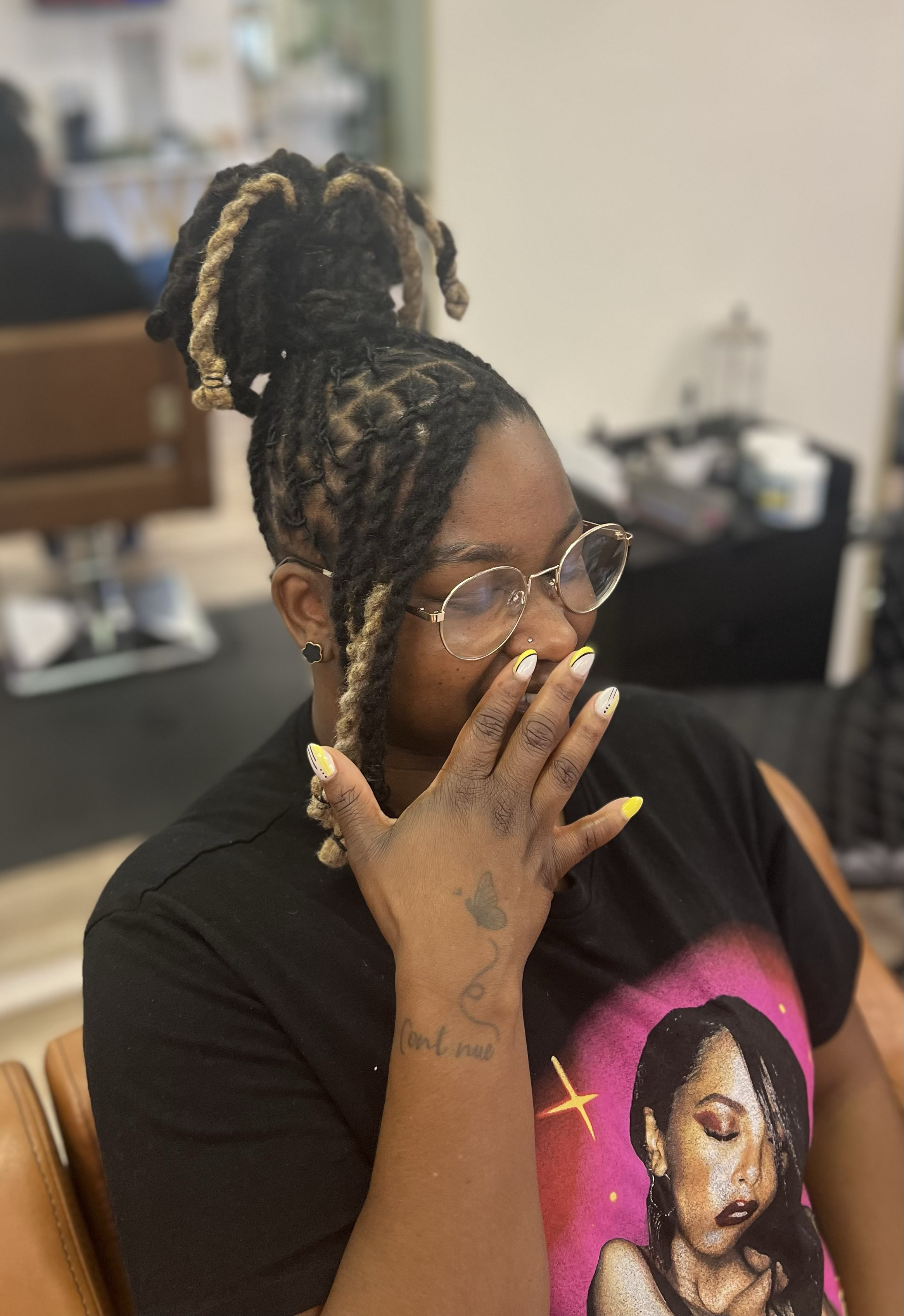 A woman with glasses and dreadlocks styled in a high bun with blonde tips, wearing a black t-shirt with a graphic of a woman and stars, covers her mouth with her hand, showing yellow and white nails and a butterfly tattoo on her arm.